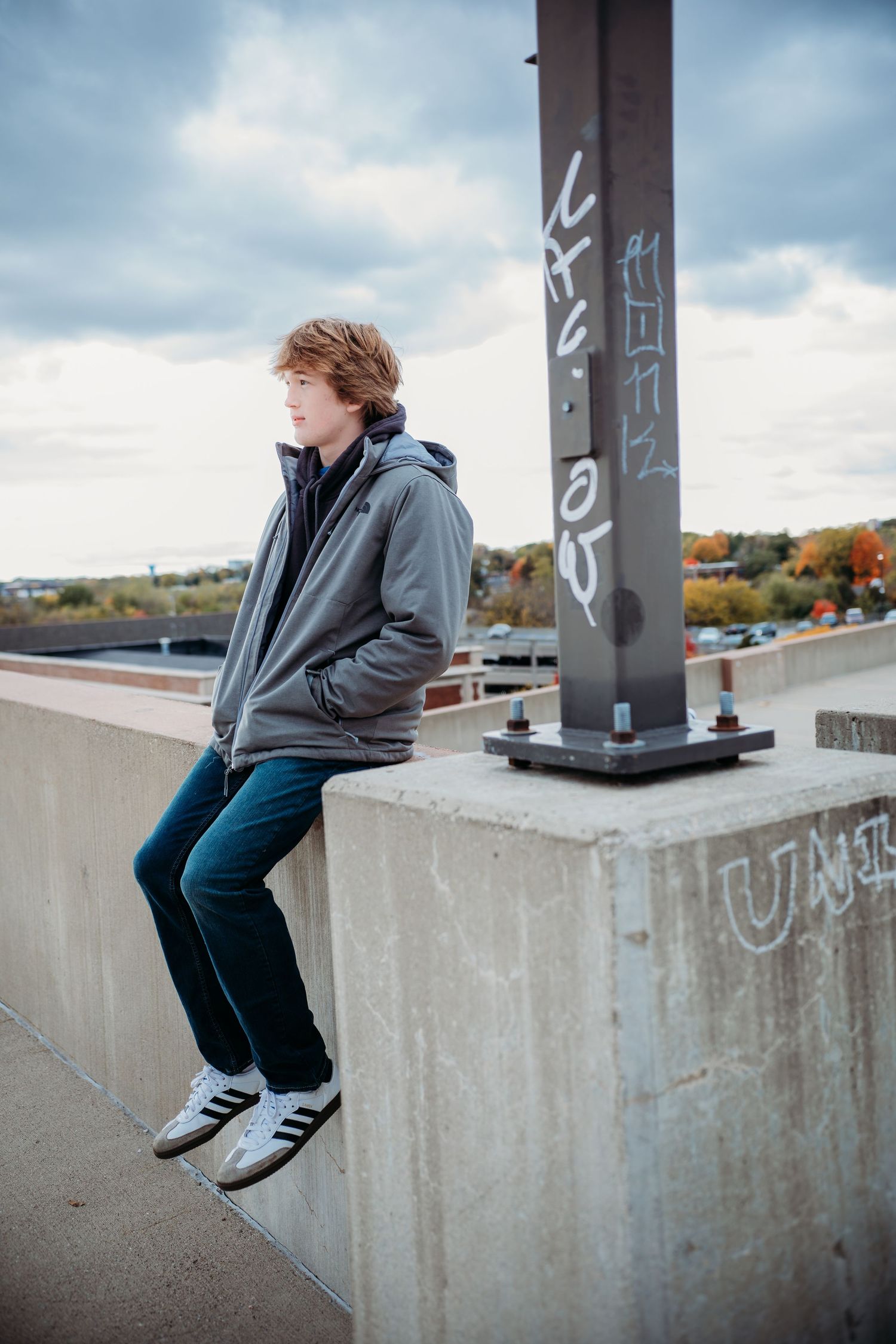 A person in a gray hoodie and jeans sits on a concrete ledge against a cloudy sky with autumn trees in the background.