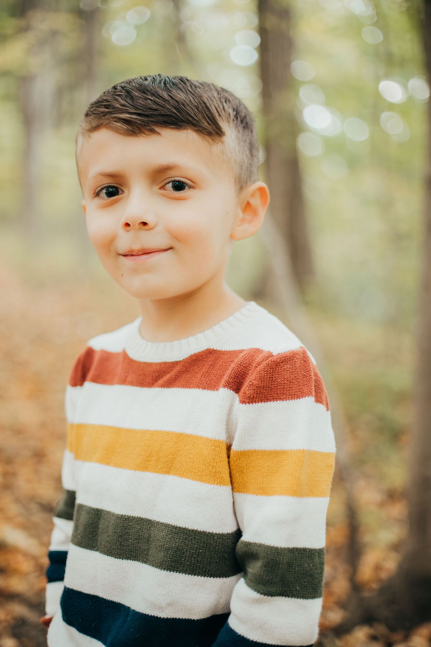 A young child in a colorful striped sweater poses outdoors in an autumn forest setting with soft bokeh lighting.