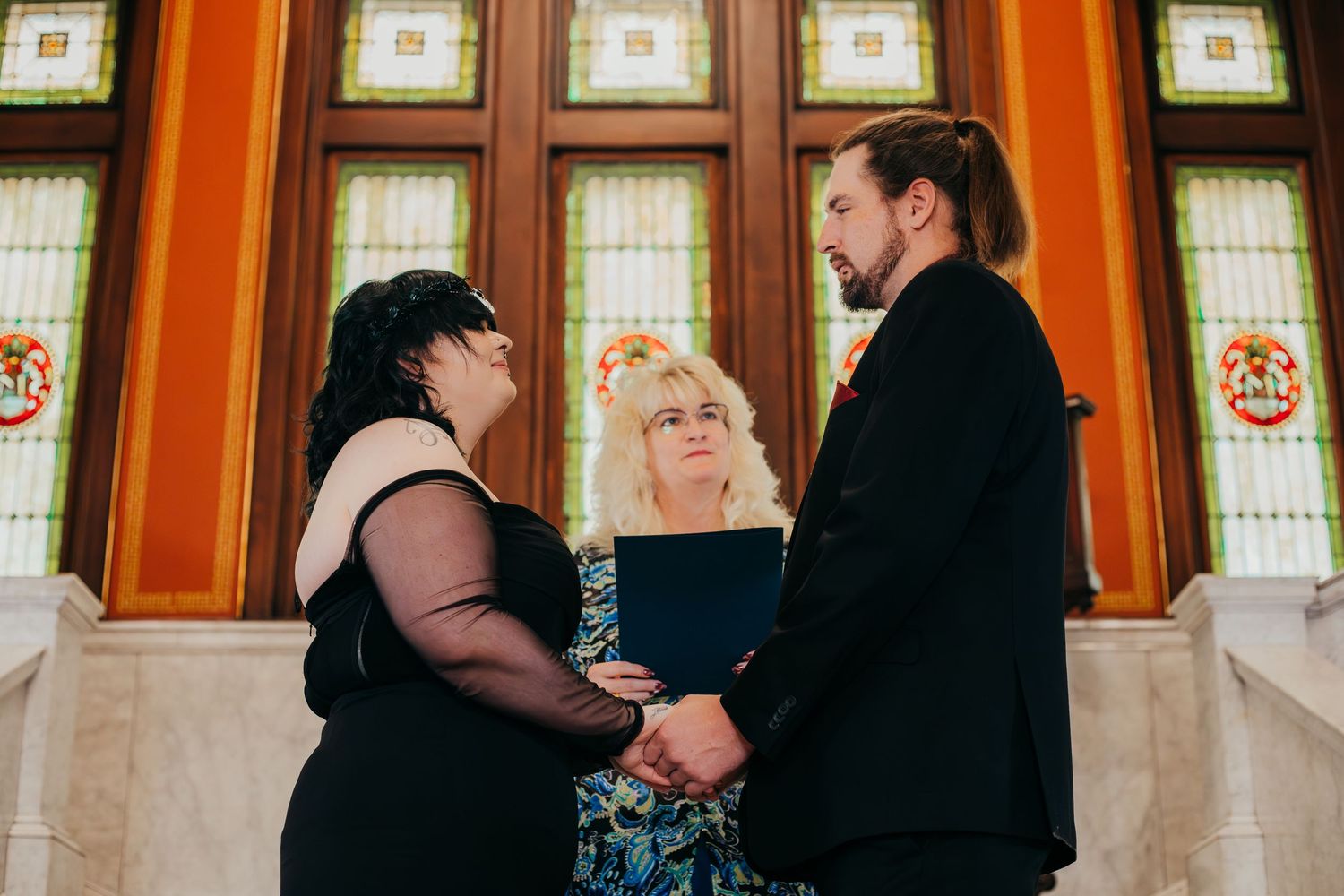 A couple holds hands during an intimate wedding ceremony with an officiant in front of stained glass windows.