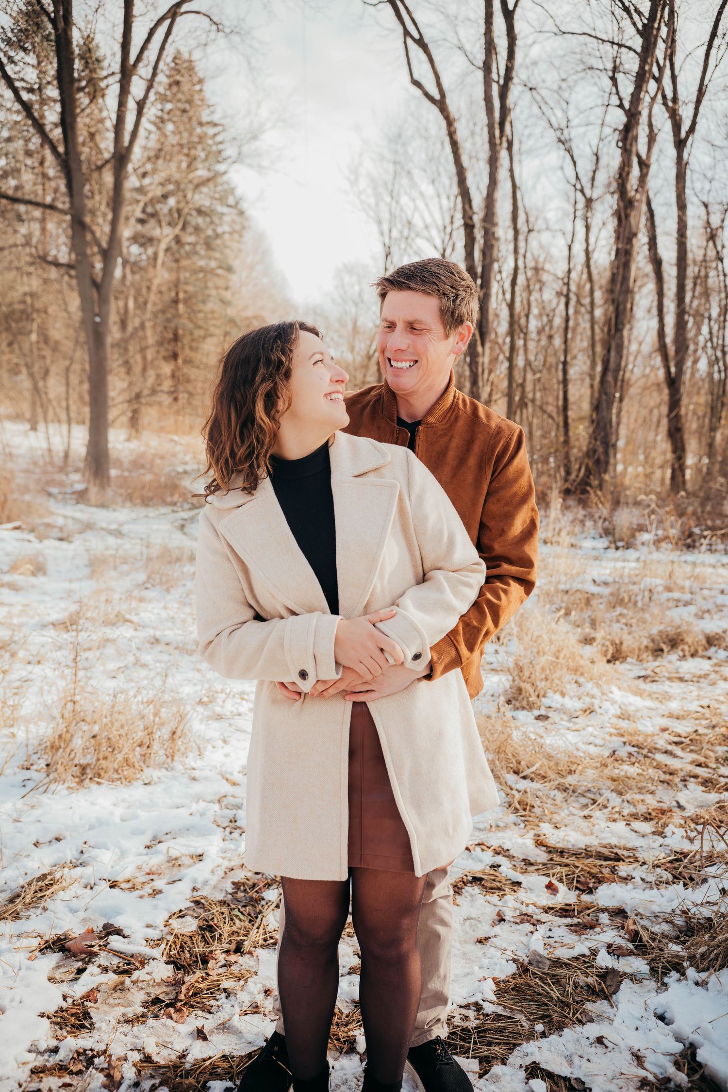 A romantic winter photoshoot in a snowy forest with a couple embracing in warm coats against a serene natural backdrop.