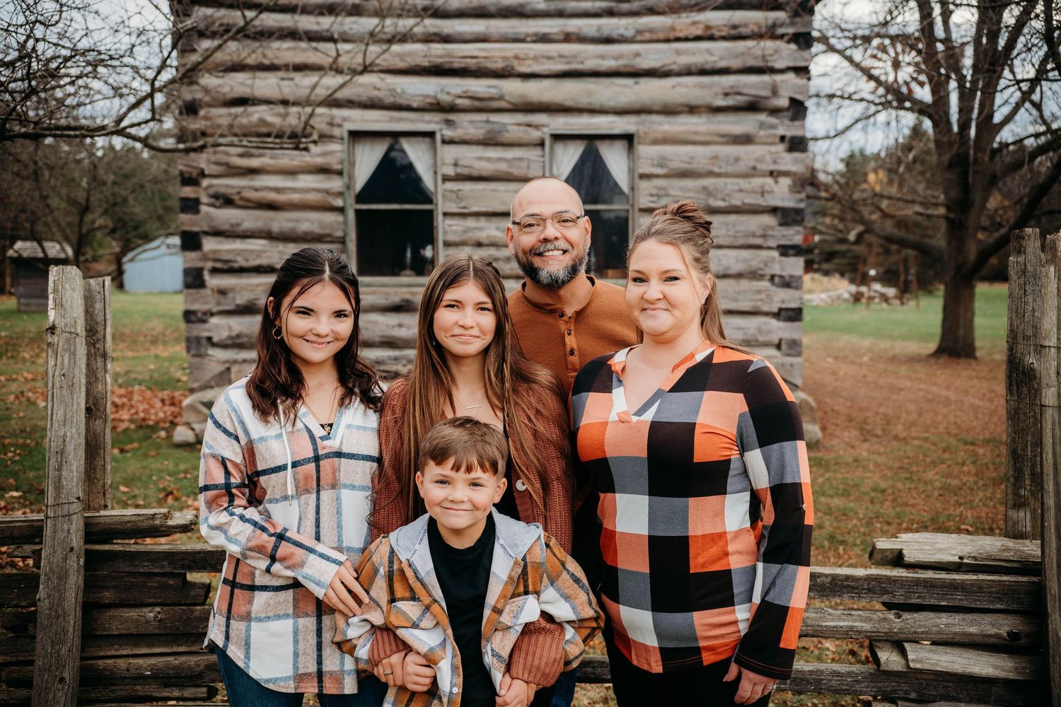 A family wearing coordinated orange and white plaid outfits poses together in front of a rustic wooden cabin.
