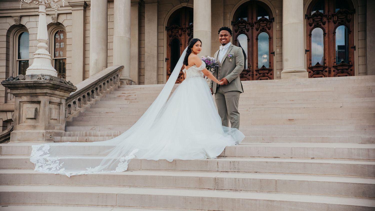 A couple in wedding attire poses on elegant stone steps of a historic building with ornate architecture.