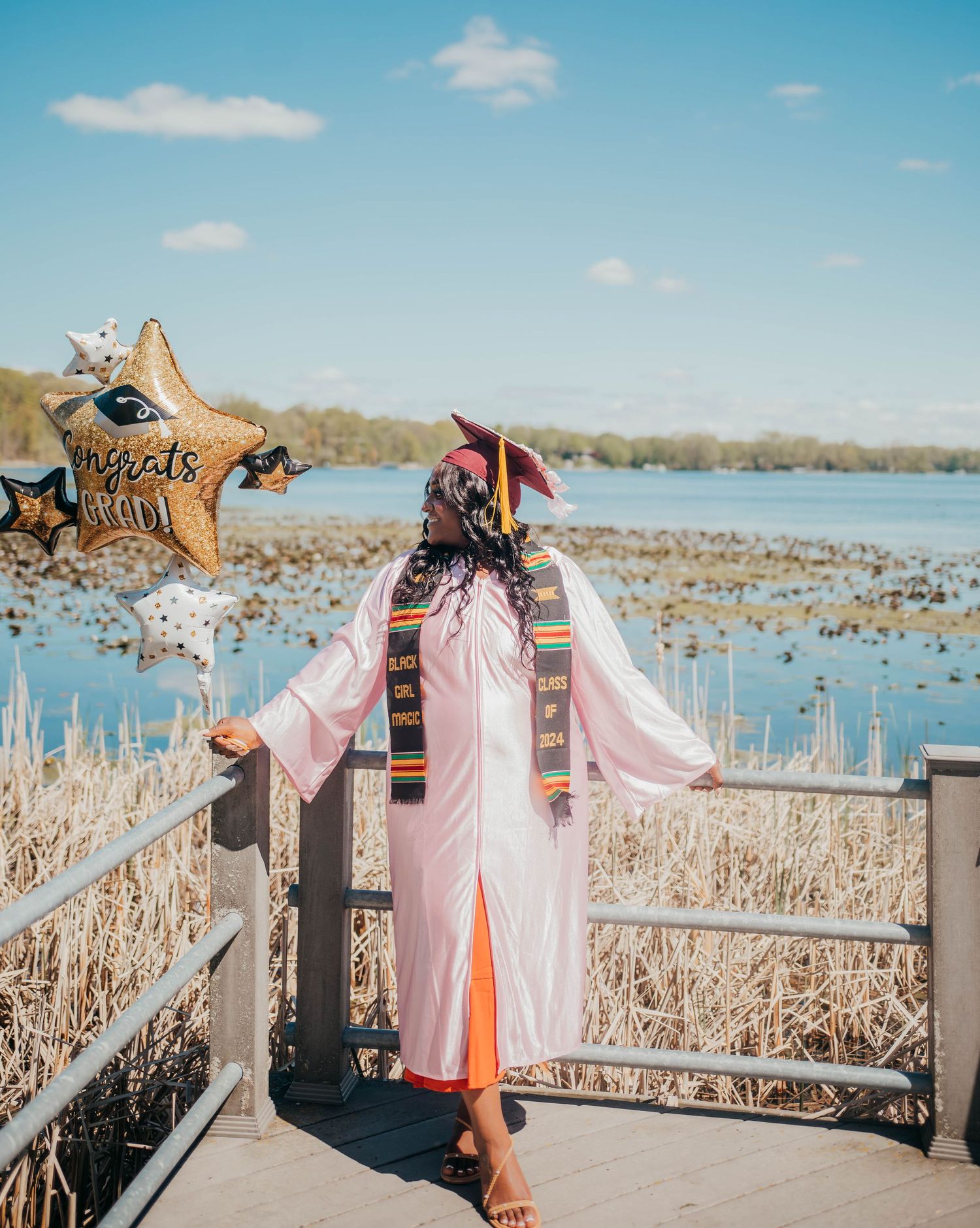 Pink dresses hanging on a rustic wooden dock railing overlooking a lake with cattails and blue sky in the background.