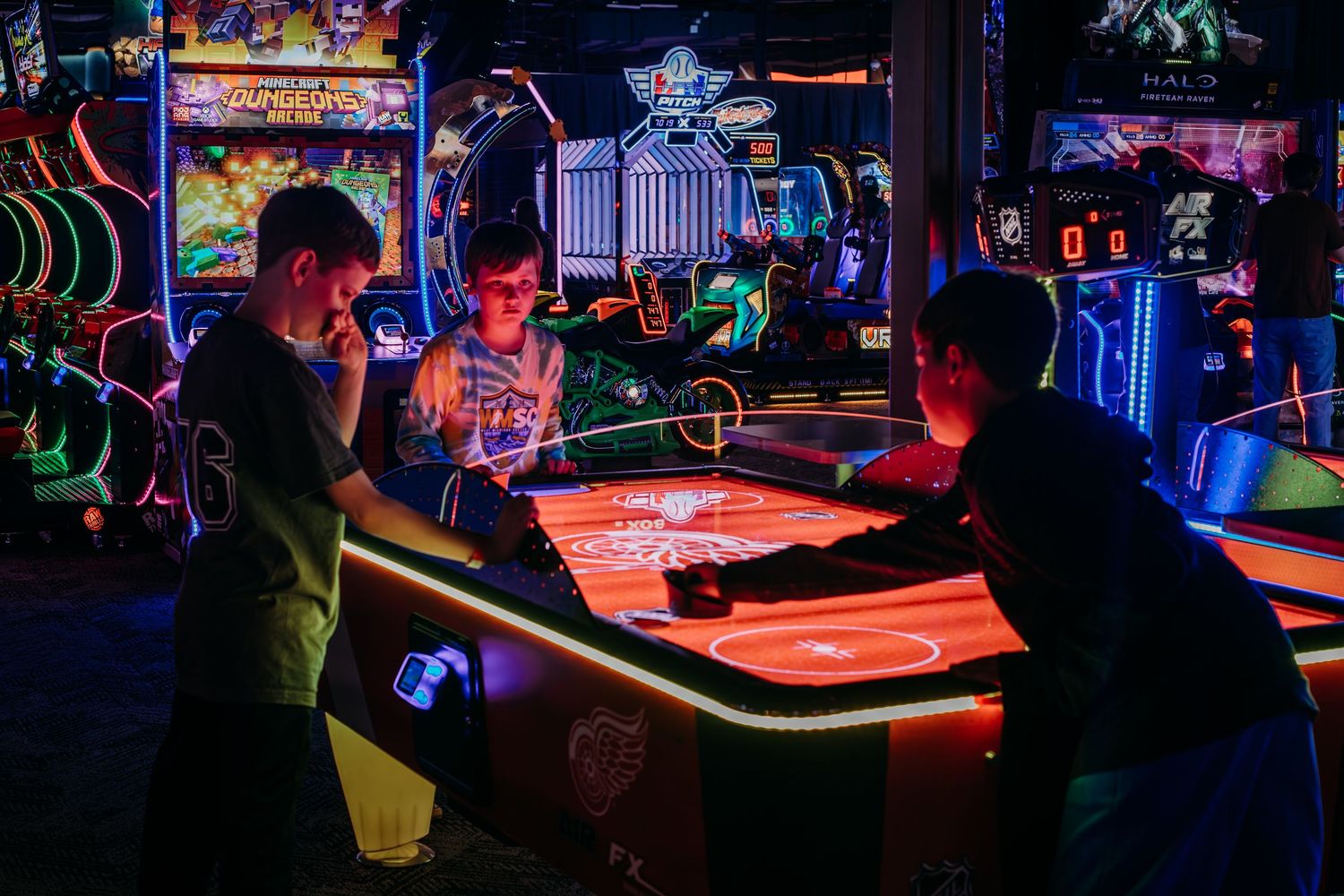 Silhouettes of people playing arcade games in a dark room illuminated by colorful neon lights and game screens.