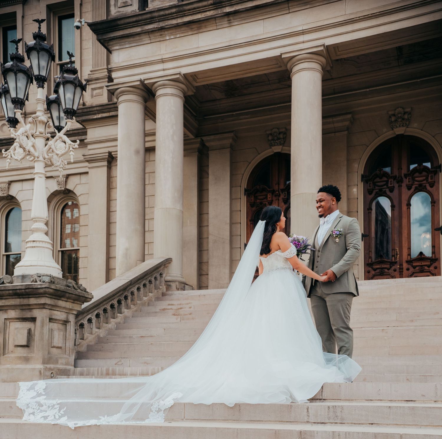 A bride in a flowing white gown poses on the grand steps of a historic stone building with ornate architecture.
