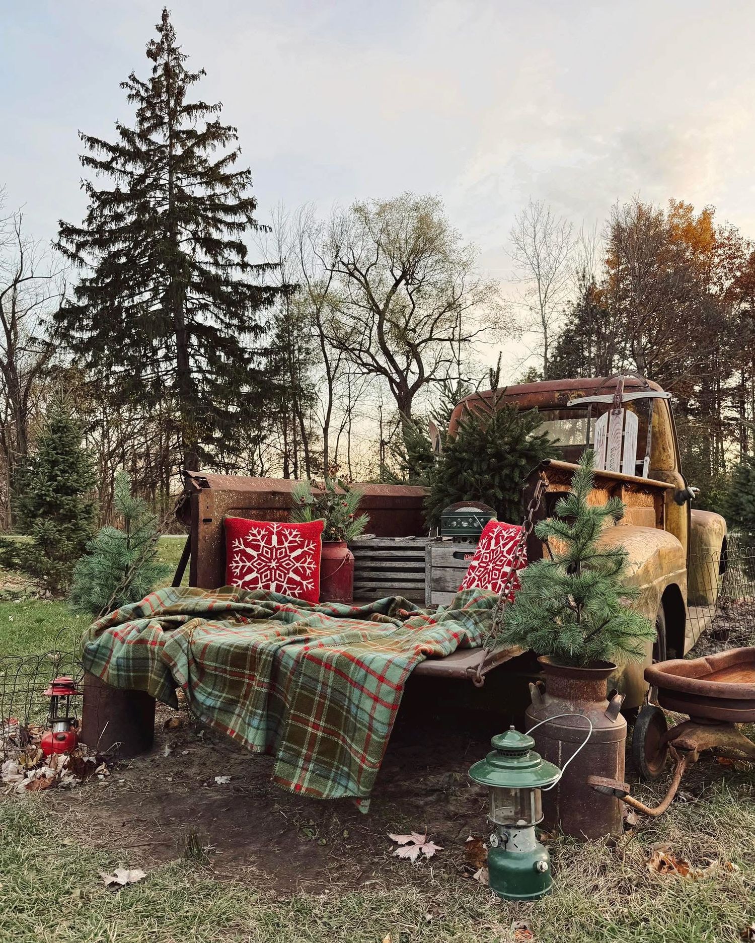A vintage truck decorated with plaid blankets, Christmas trees, and red holiday decor creates a rustic outdoor holiday display.
