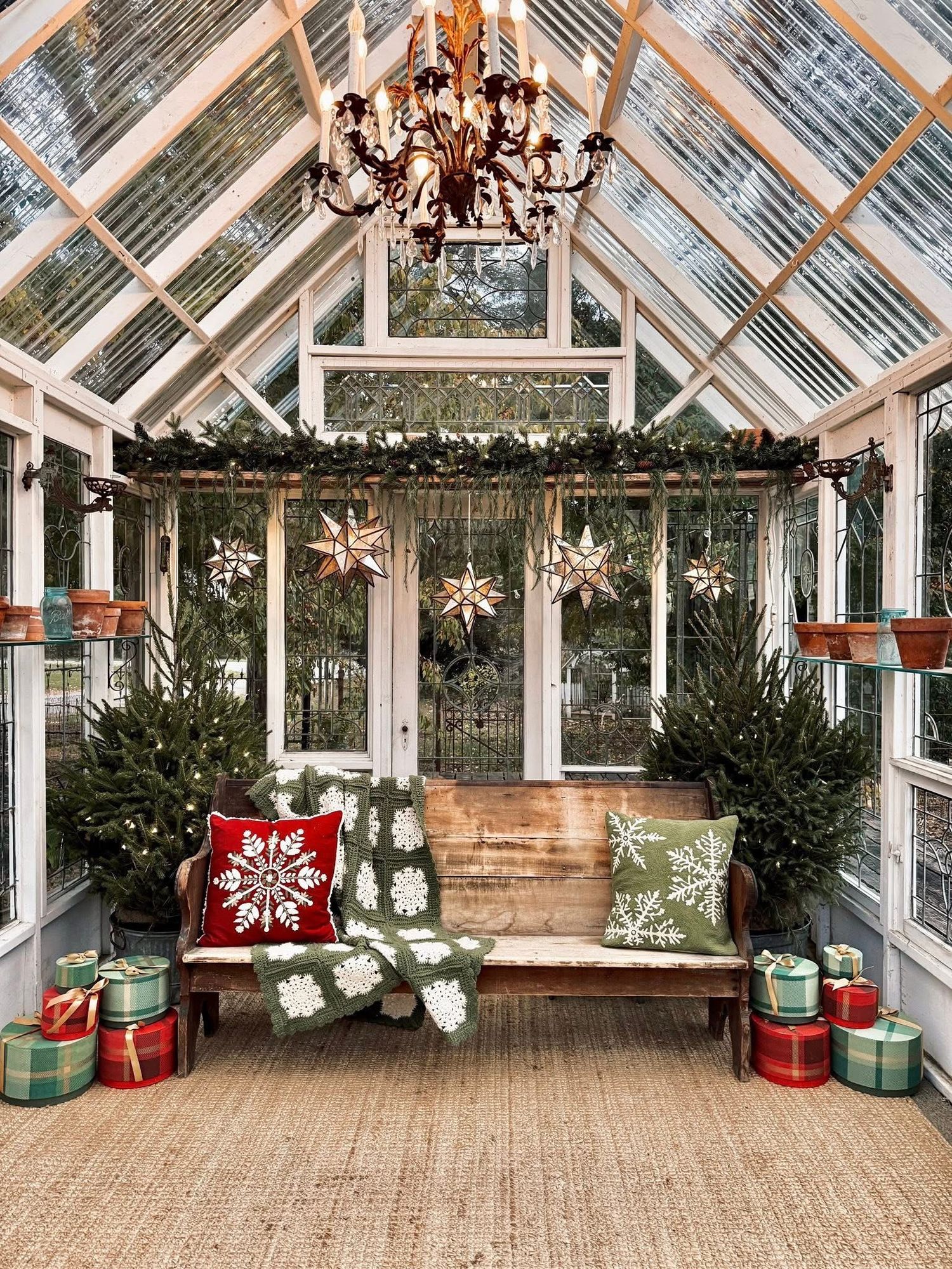 A bright greenhouse sunroom decorated for winter with evergreen trees, red pillows, and hanging snowflake ornaments.