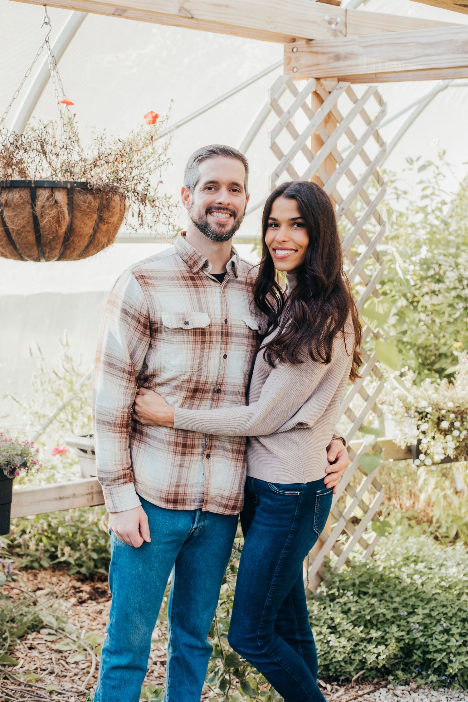 A couple wearing matching plaid shirts and jeans embrace while standing in a greenhouse with natural light.