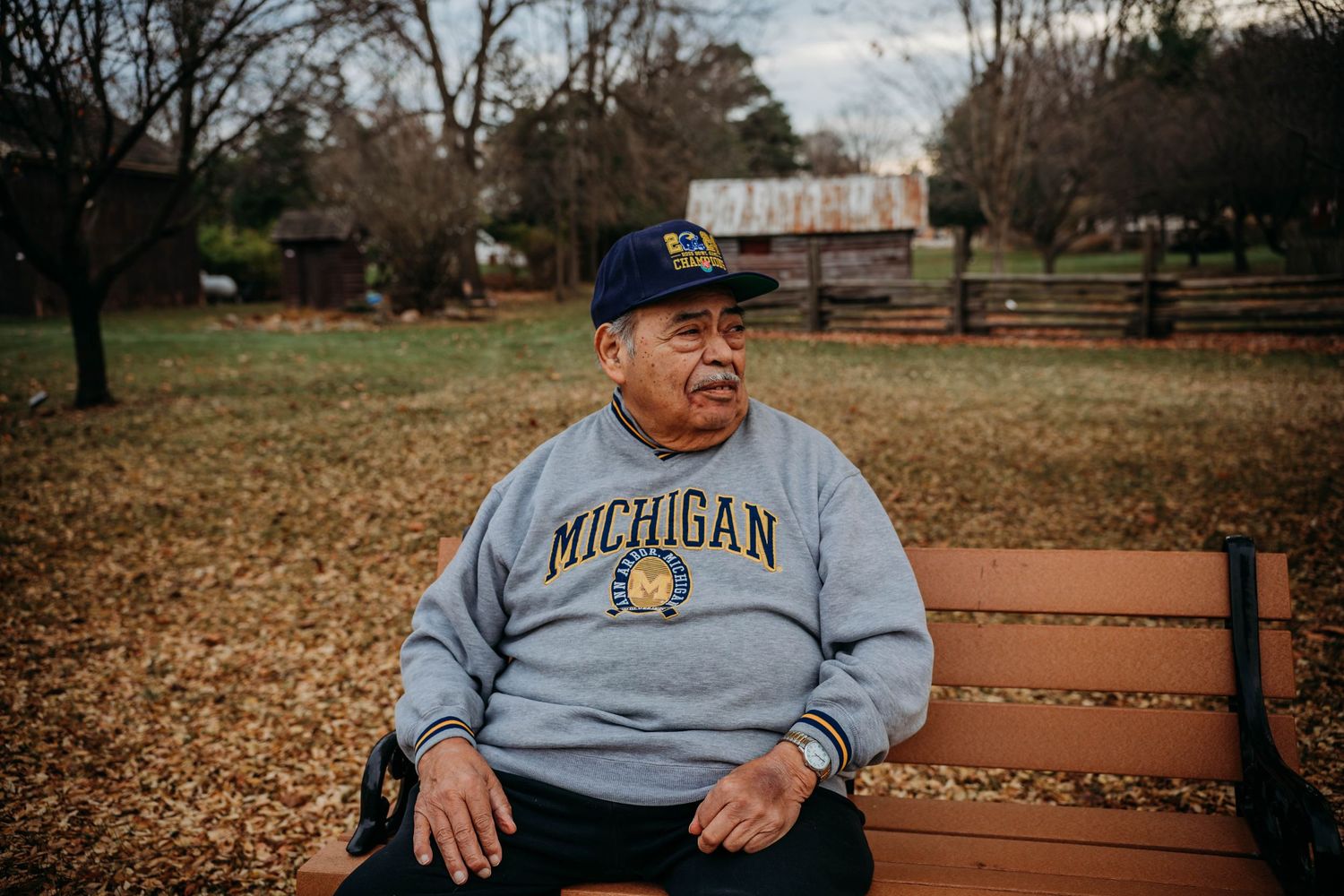 A person wearing a Michigan sweatshirt sits on a wooden bench in an autumn park setting.