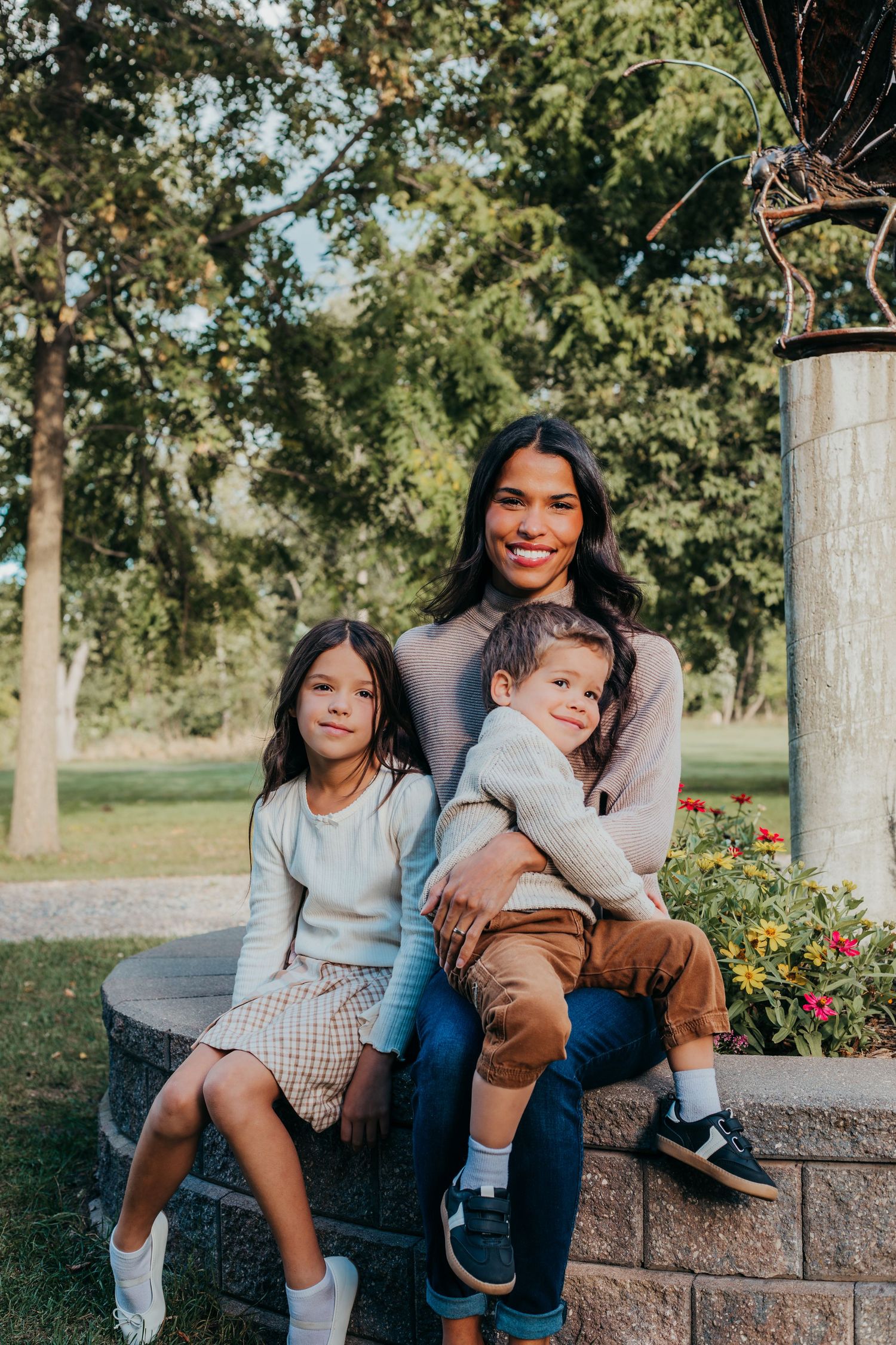 Family sitting together on stone wall in park during autumn with trees and flowers in background.