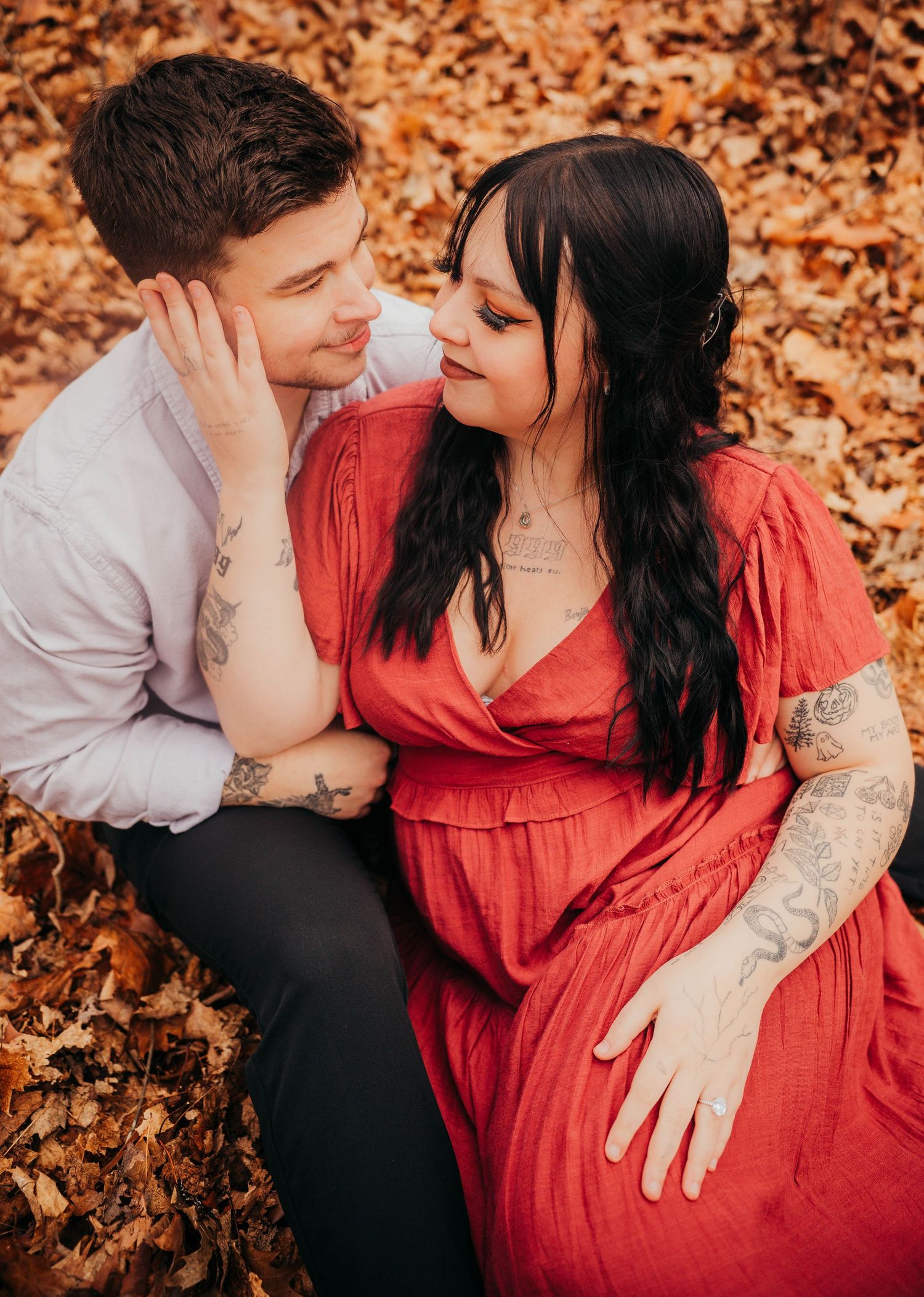 Romantic couple sitting on autumn leaves with woman in red dress and man in white shirt sharing intimate moment.