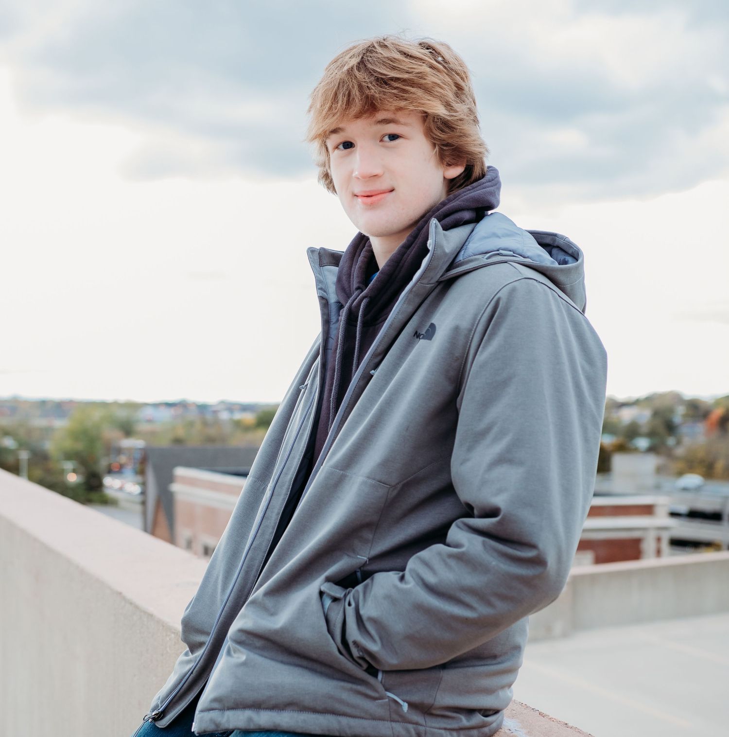 Person in gray hoodie sitting outdoors against concrete wall with urban backdrop on overcast day.