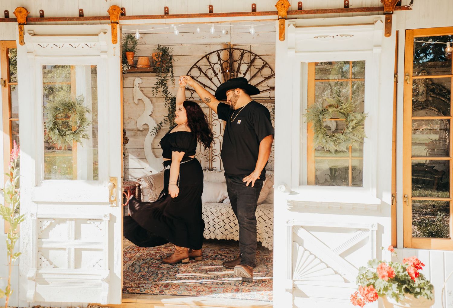 Two people in black outfits pose together on a rustic farmhouse porch with white vintage doors and floral decor.