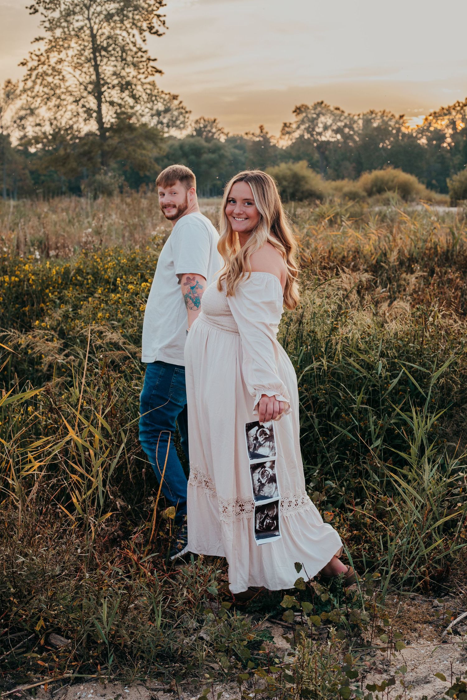 A couple embraces in a golden field at sunset holding ultrasound photos during a pregnancy announcement photoshoot.