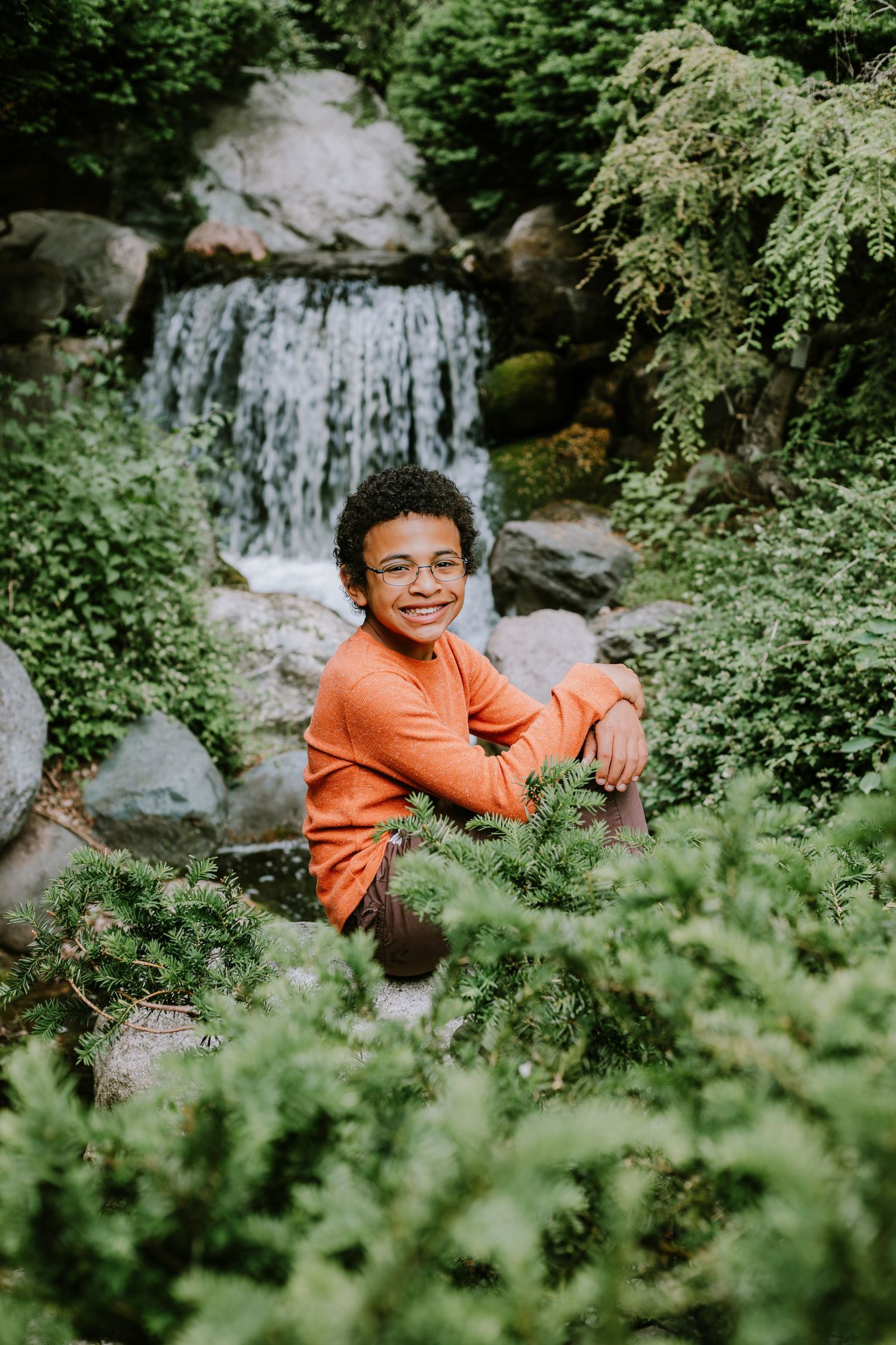 A person in an orange shirt sits among lush greenery with a cascading waterfall visible in the background.