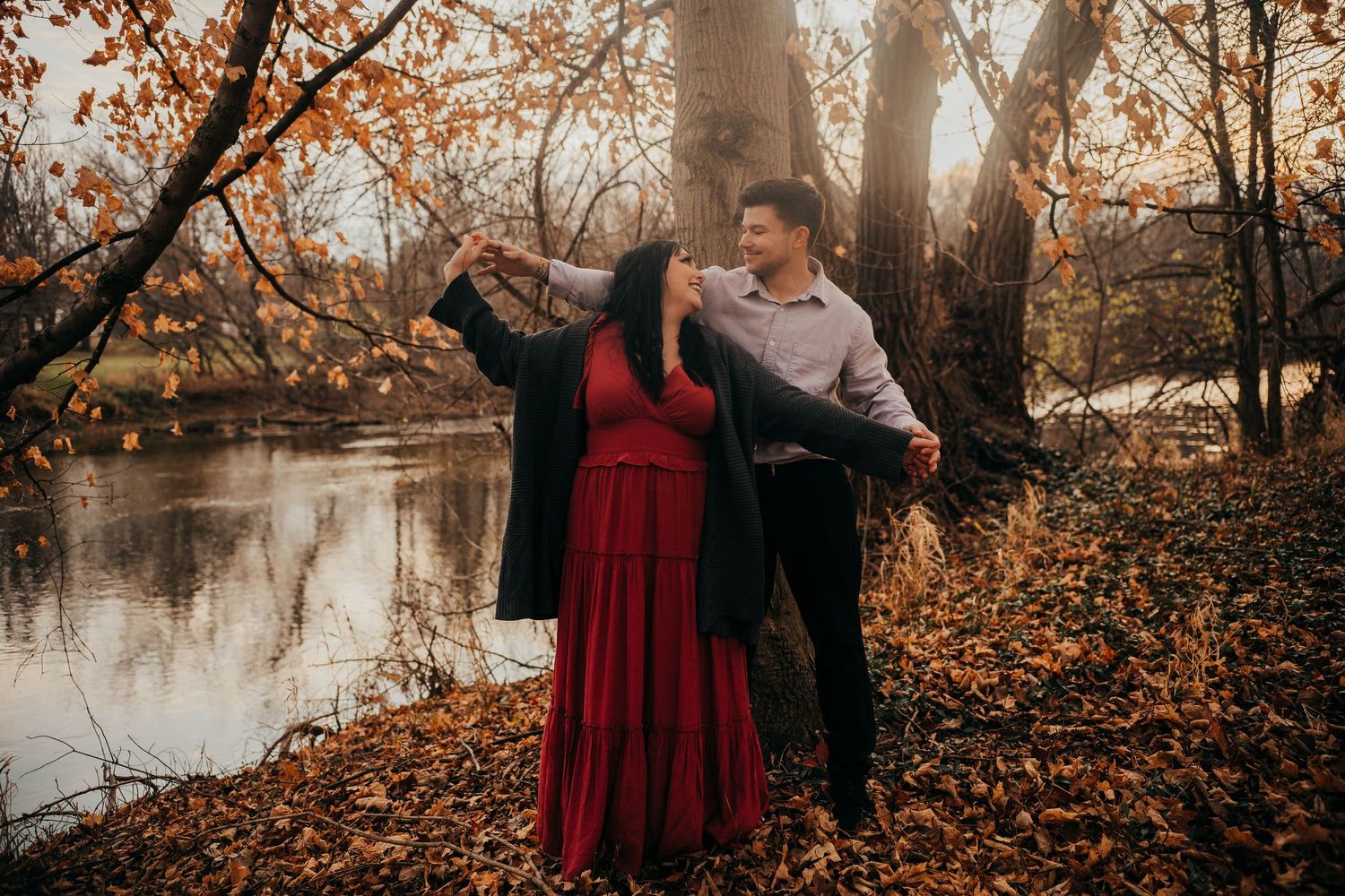 A couple in autumn attire poses romantically by a tranquil lake surrounded by fall foliage at sunset.