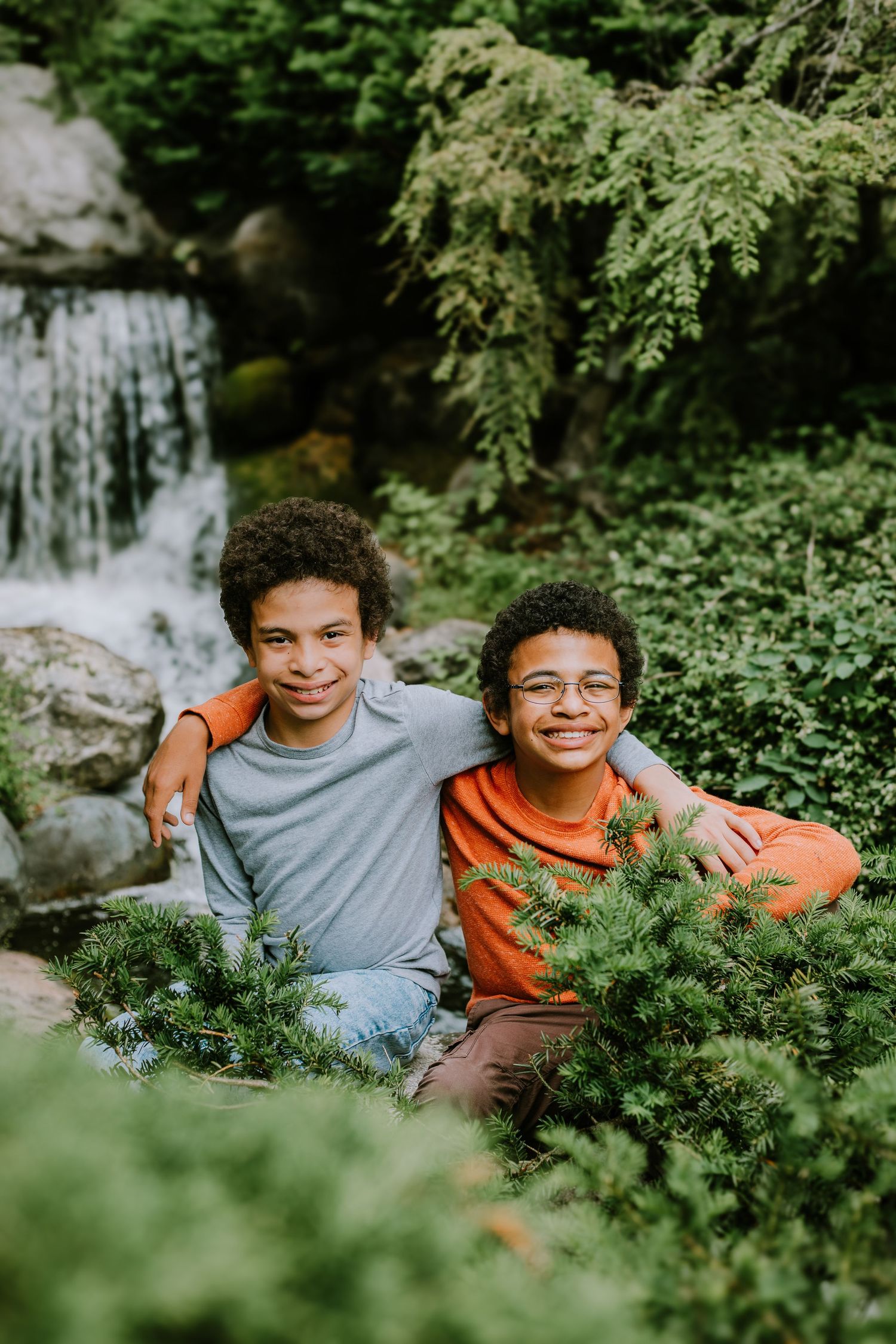 Two people sit together outdoors surrounded by lush green foliage and a waterfall in the background.