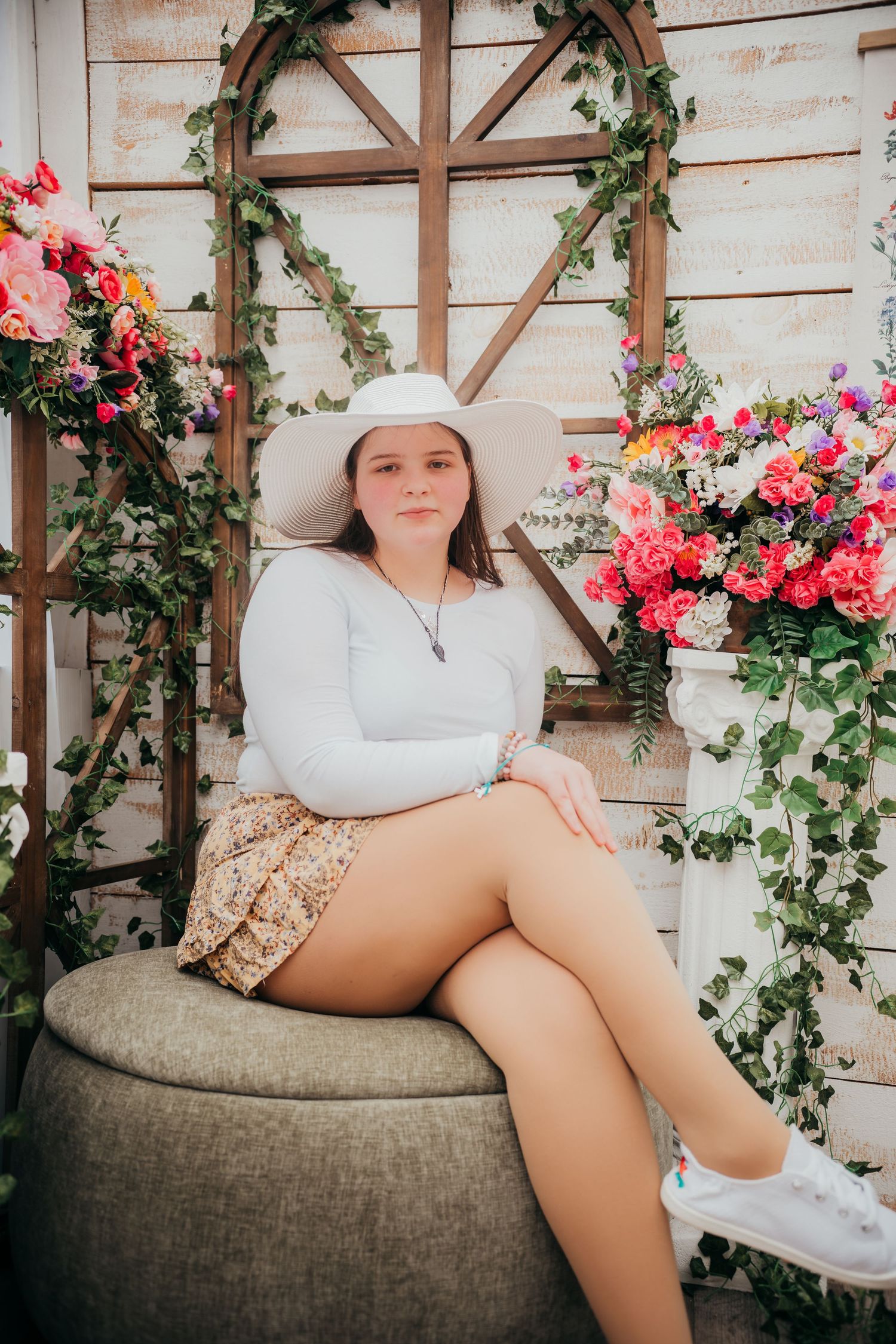 A rustic photo setting with wooden panels, floral decorations and someone seated on a gray ottoman wearing a white hat.
