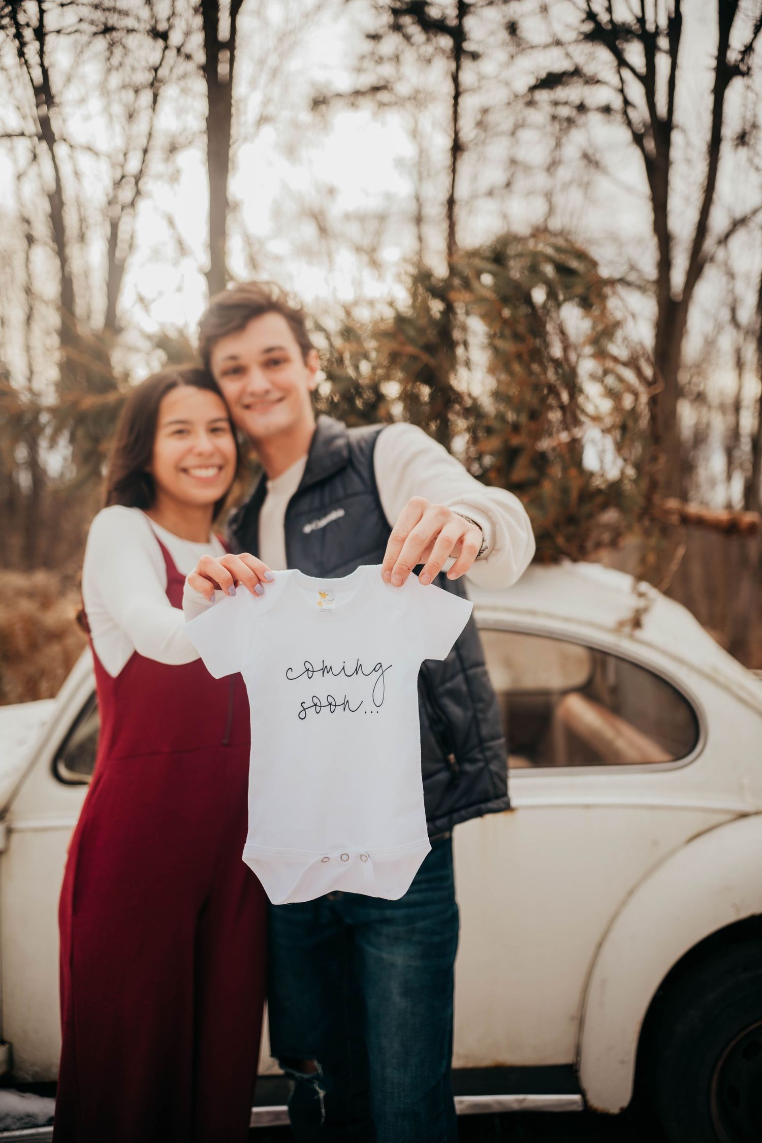 A couple holds up a small white baby onesie while standing in front of a vintage Volkswagen Beetle on an autumn day.