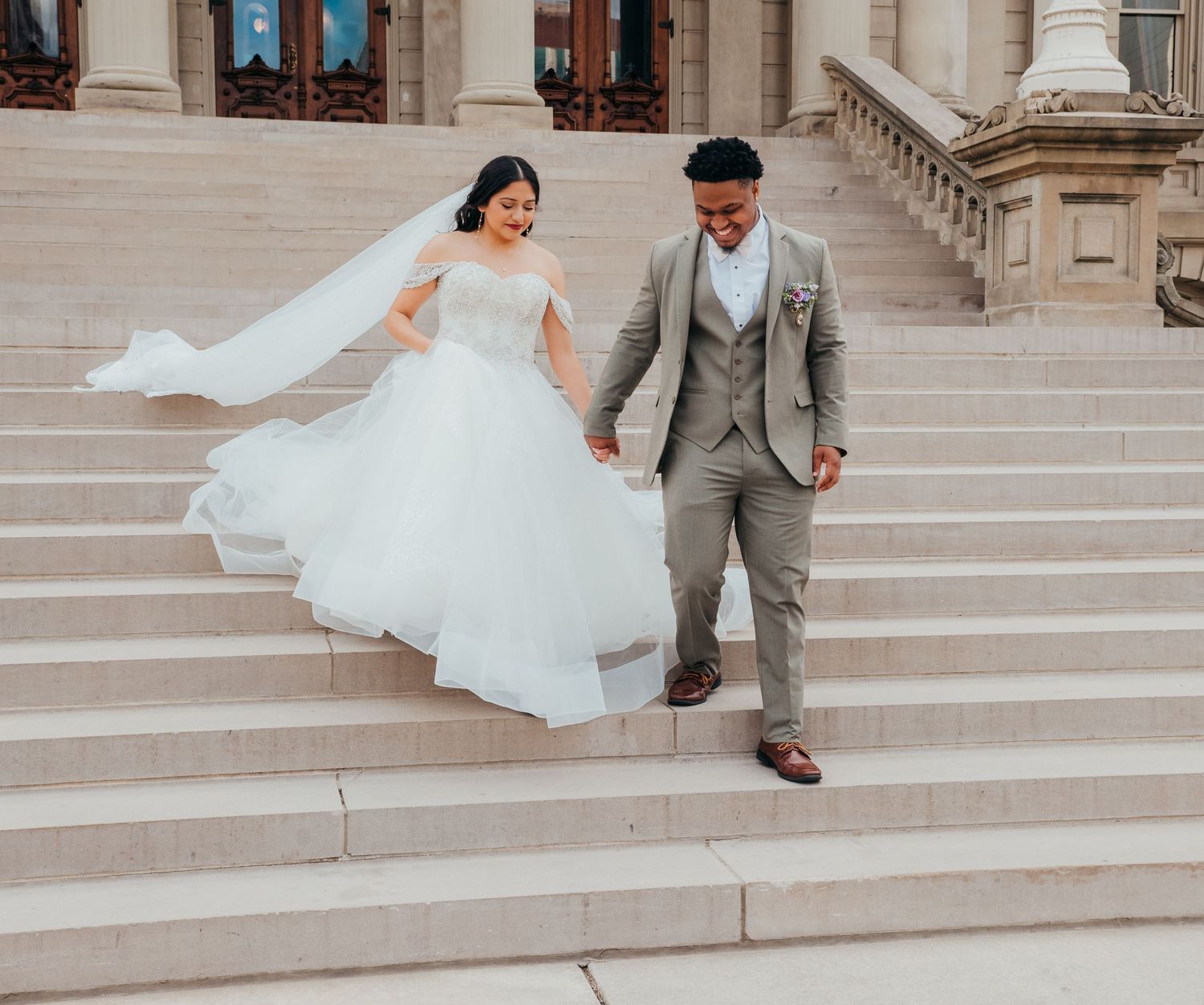 A couple descends elegant stone steps, with the bride's white veil and gown flowing in the breeze as they hold hands.