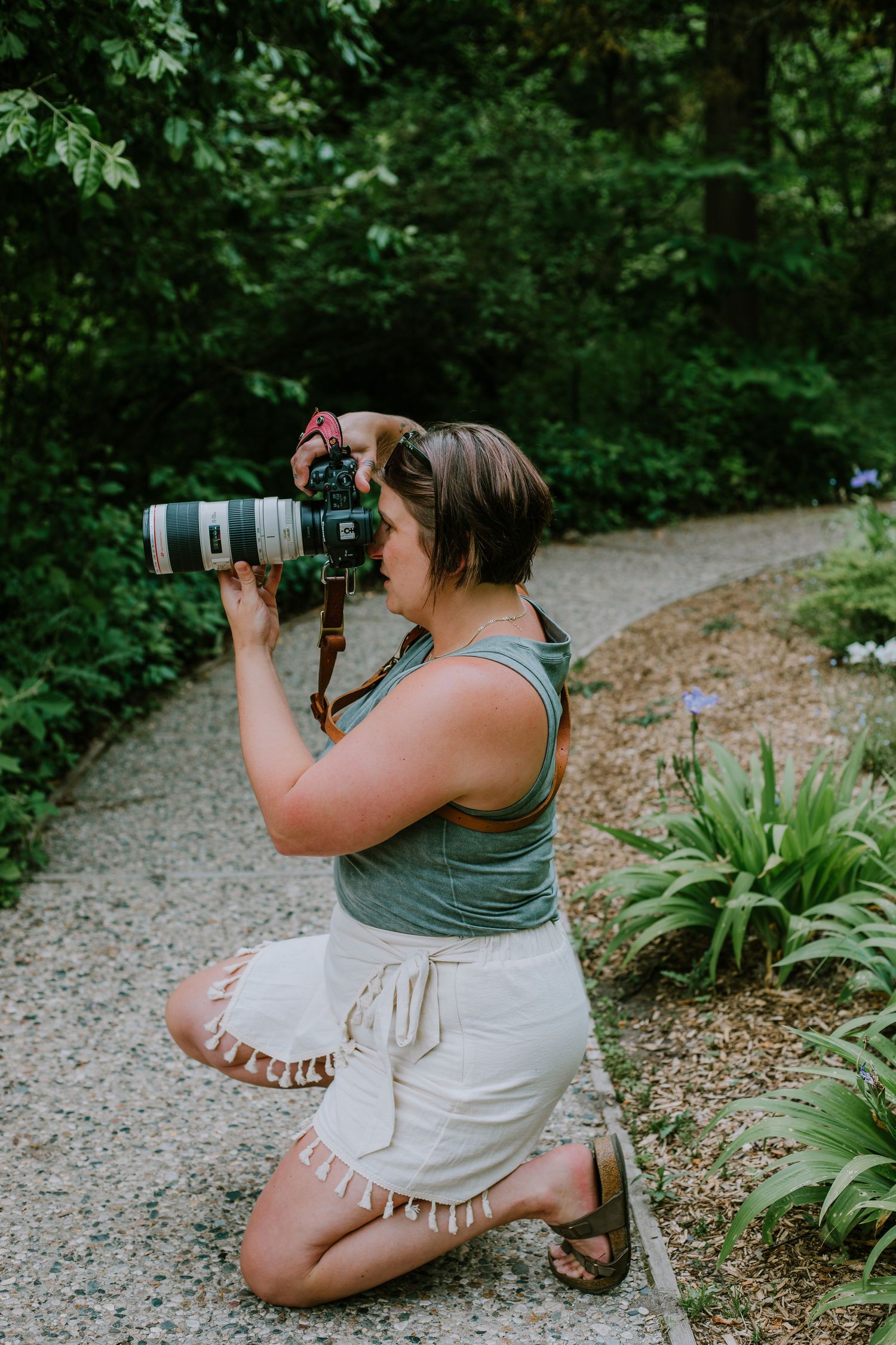 A photographer in a tank top and white shorts kneels on a gravel path while taking photos with a telephoto lens.