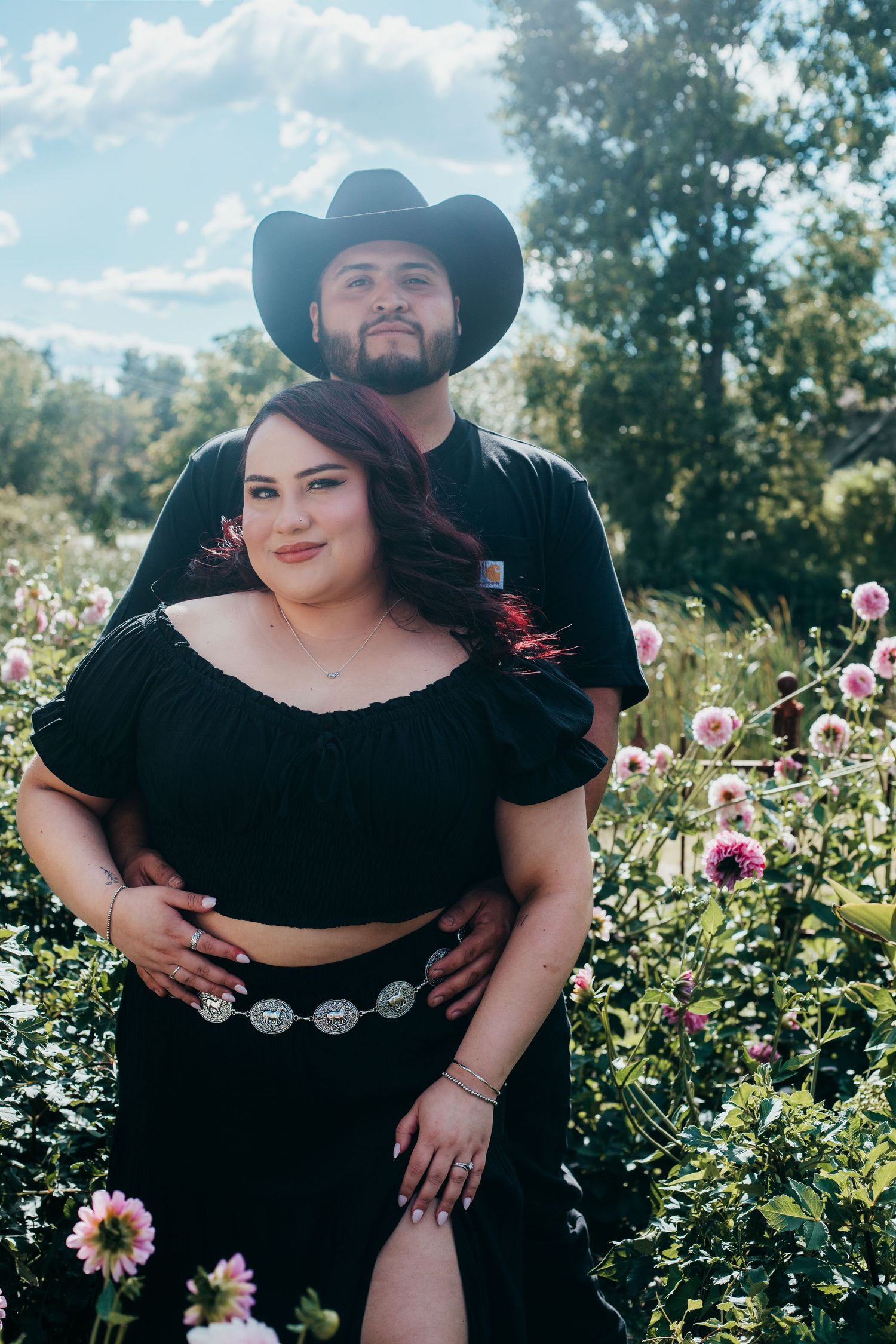Couple in black western attire embraces in a garden filled with pink and white flowers on a sunny day.