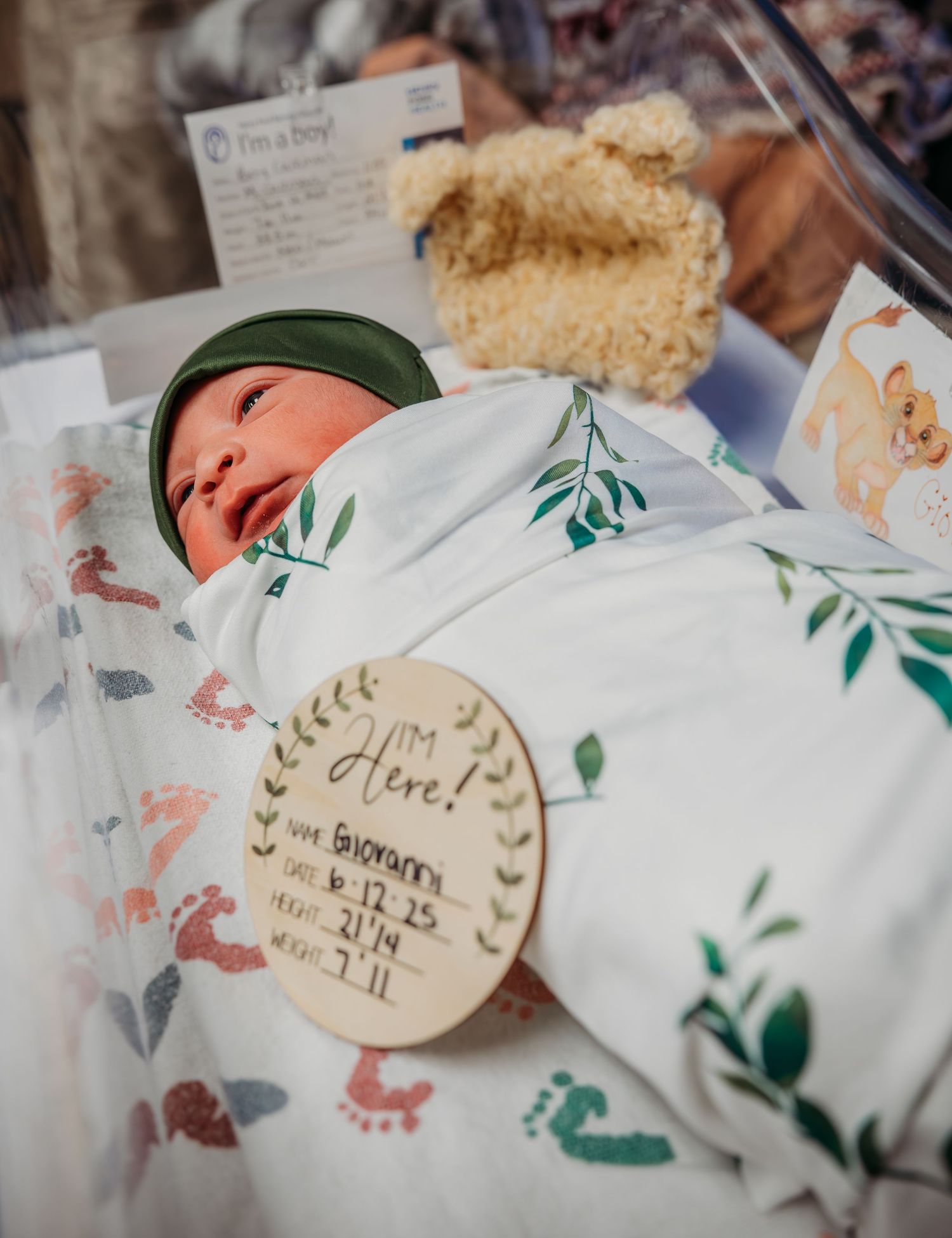 A newborn baby rests in a hospital bassinet with a wooden birth stats sign and floral print swaddle blanket.