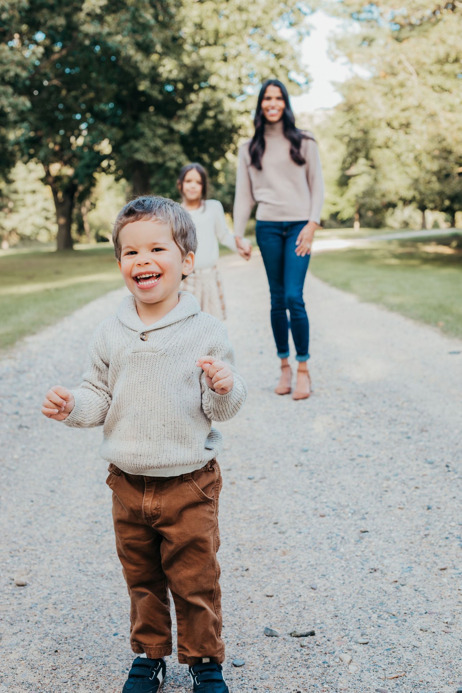 A child wearing a beige sweater and brown pants smiles joyfully while walking on a gravel path in a park setting.