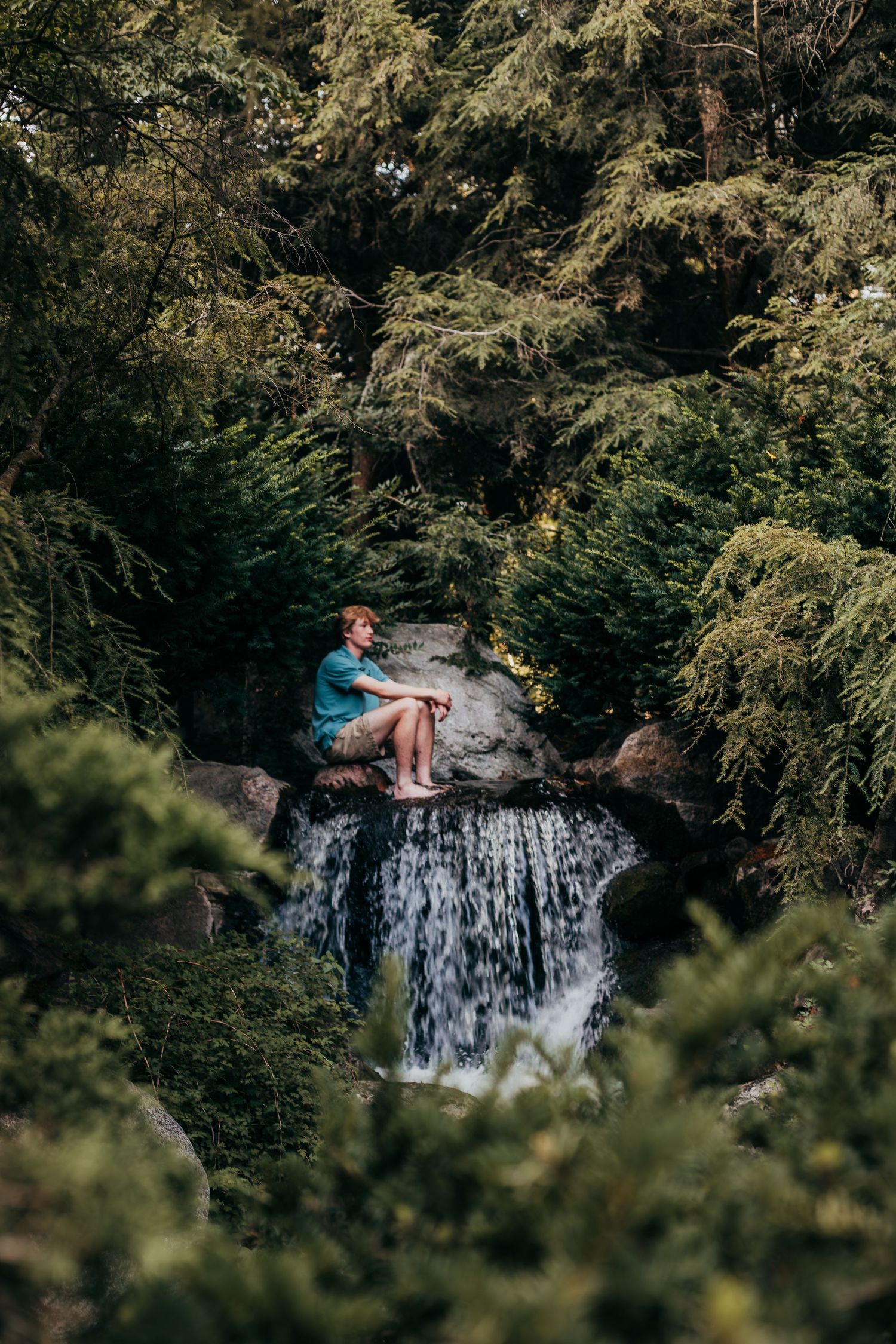 A person holding fabric stands near a cascading waterfall surrounded by lush evergreen forest in a scenic nature setting.
