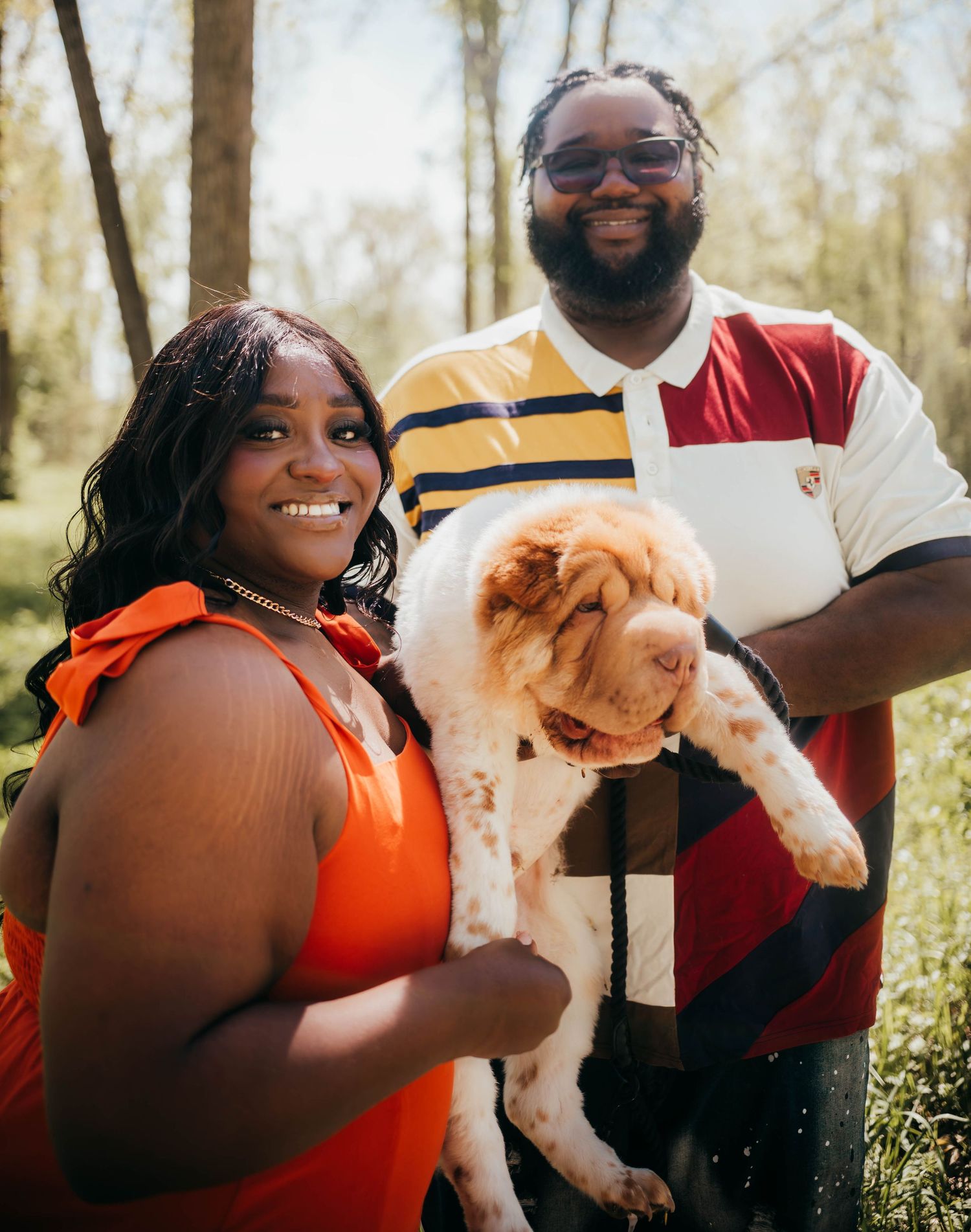 A couple poses together outdoors in coordinated outfits of orange and white while holding a small brown and white dog.