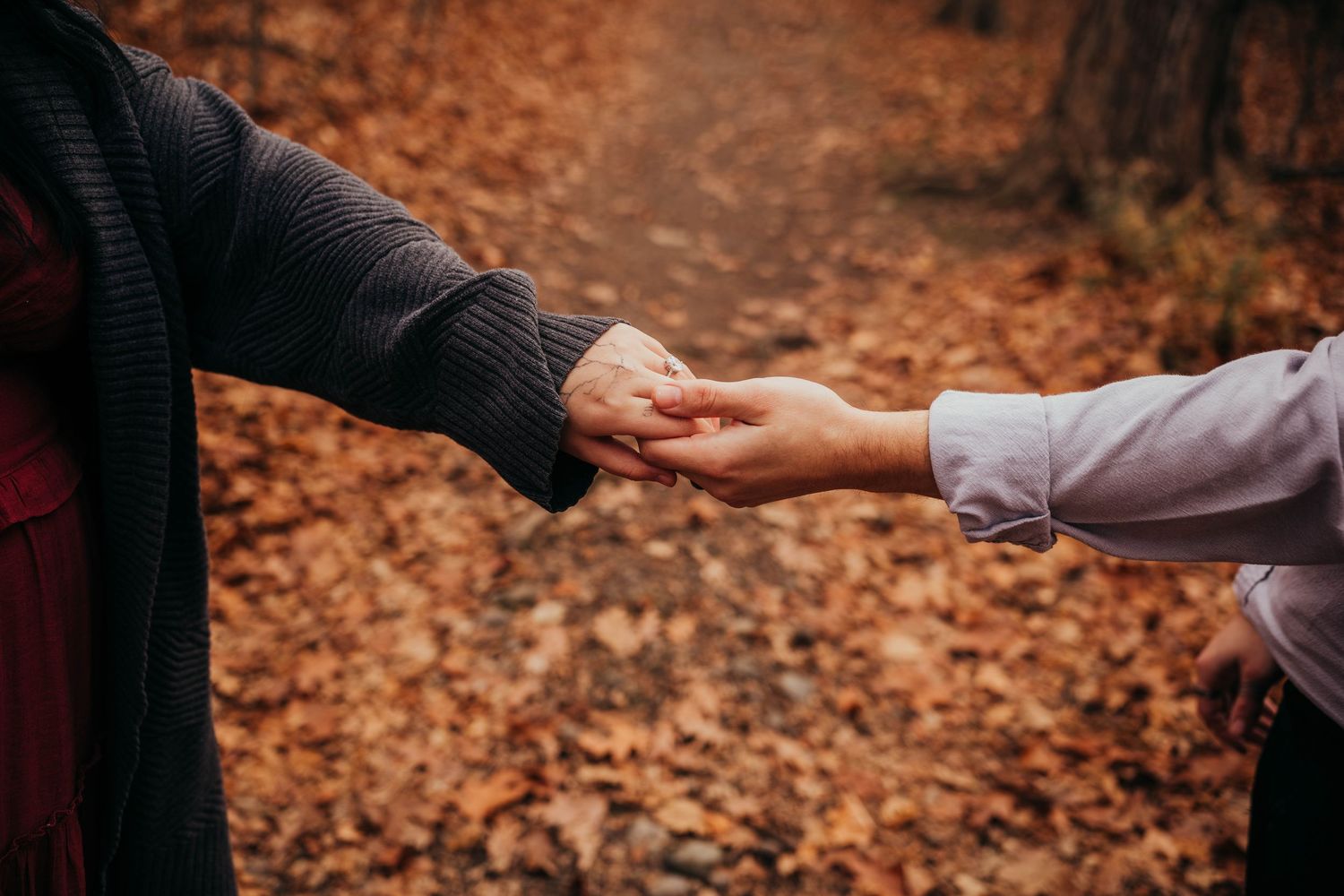 Couple holding hands while walking through autumn forest with orange fallen leaves on ground.