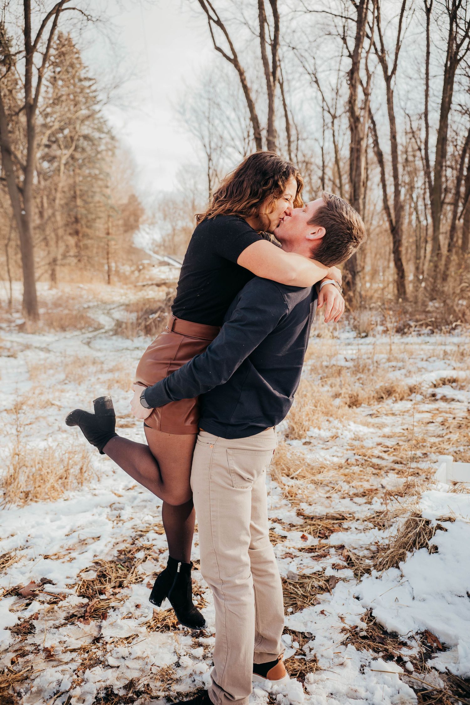 A romantic couple embraces in a snowy winter forest setting with bare trees in the background.