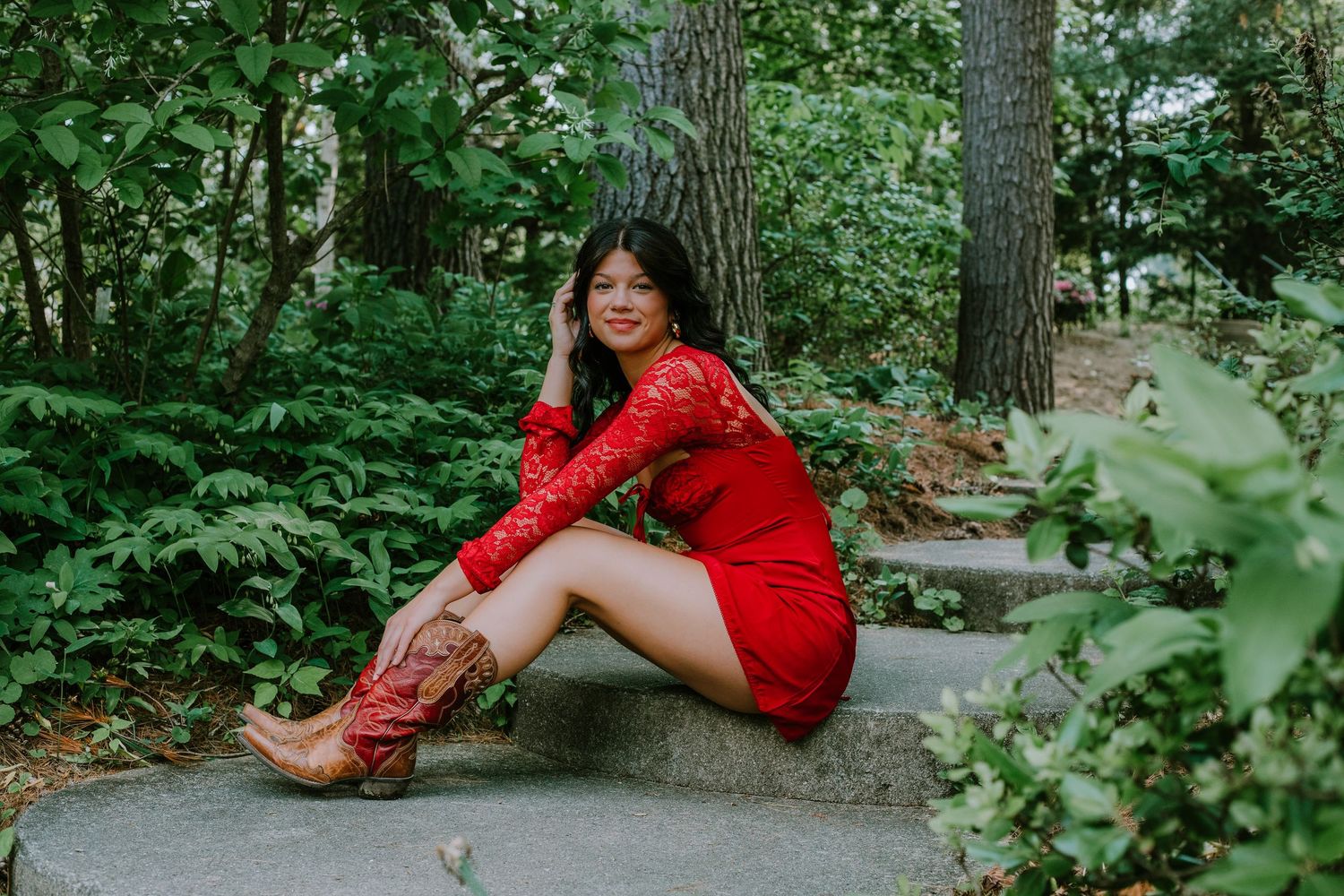 A person in a bright red dress poses gracefully on a stone path surrounded by lush green foliage in a garden setting.