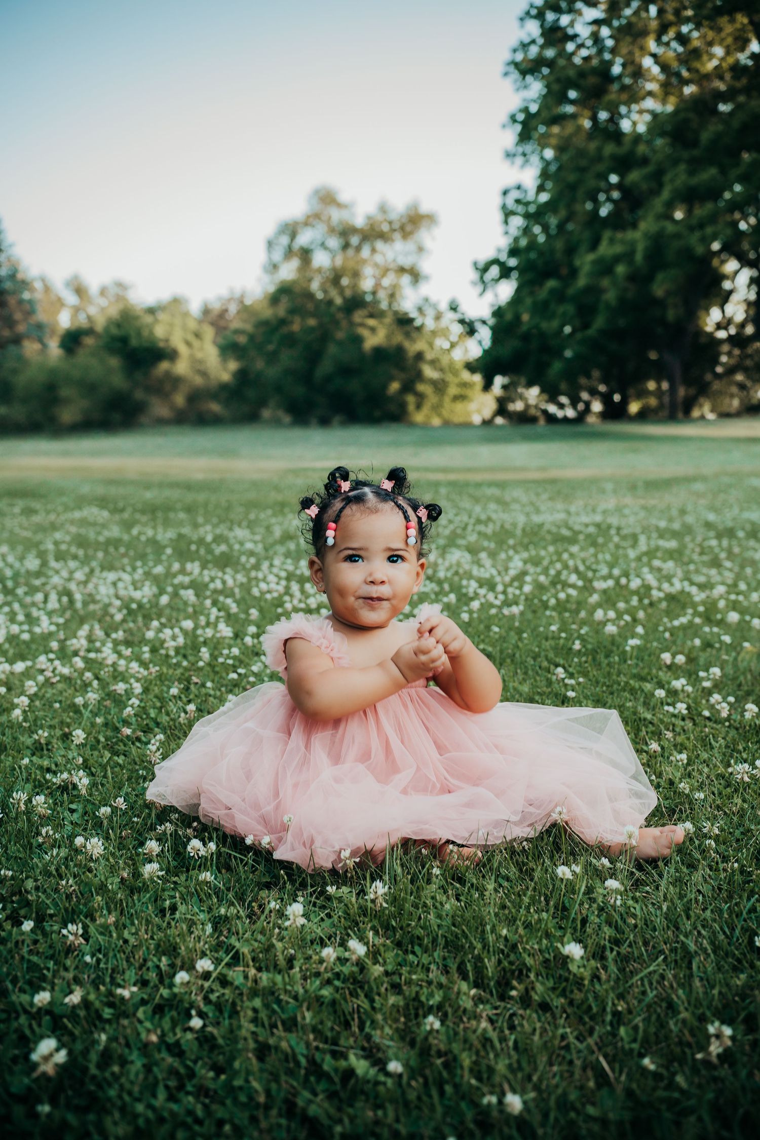 A small child in a pink tulle dress sits in a field of white wildflowers during a sunny outdoor photo session.