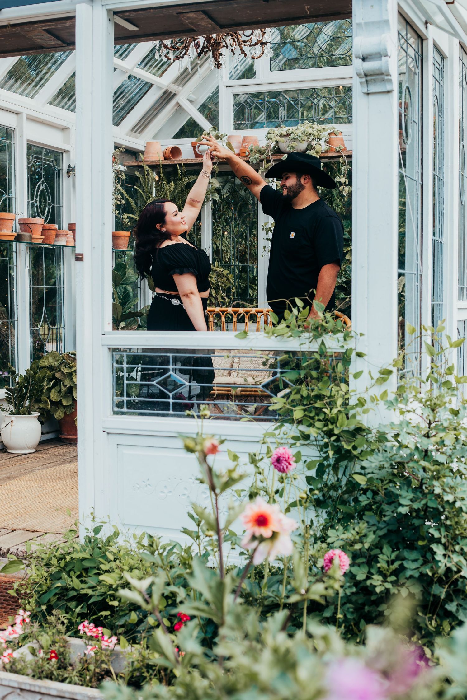 Romantic couple poses in a white greenhouse conservatory surrounded by blooming flowers and greenery.