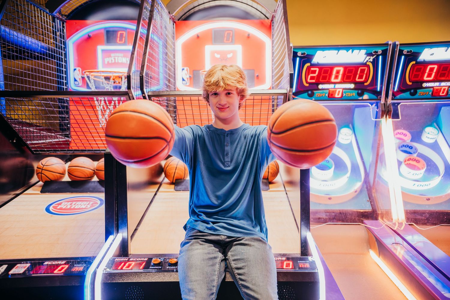 Person playing basketball arcade game with neon lights and multiple game machines in entertainment center.