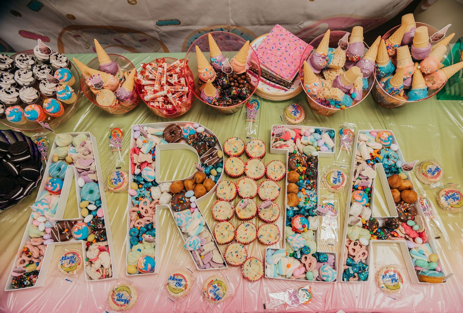 Colorful cupcakes and desserts arranged to spell out ANNA with unicorn-themed decorations on a pink tablecloth.