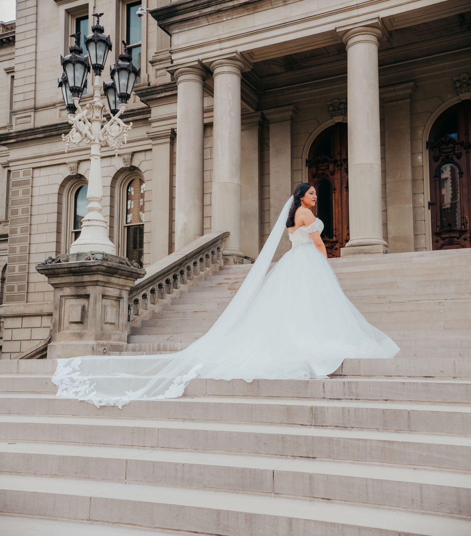 A bride in a flowing white ballgown poses gracefully on grand stone steps of a historic building.