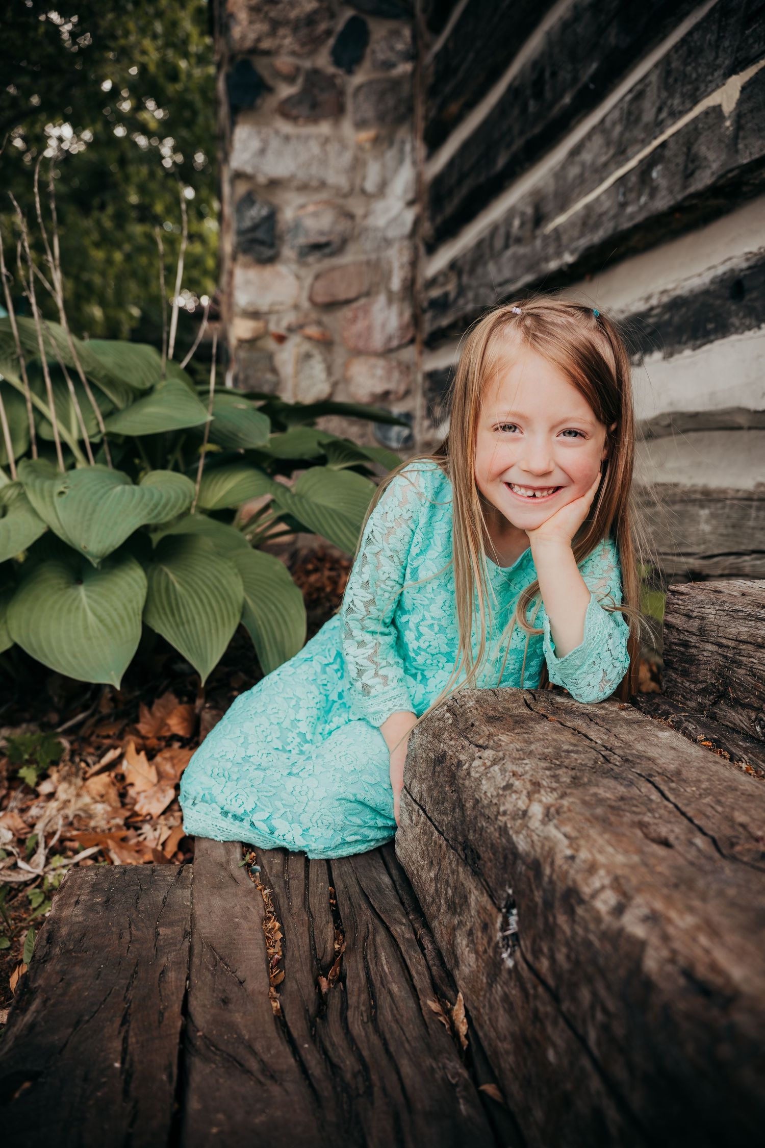Young person in a turquoise lace dress sits on rustic wooden steps next to green hostas and stone wall.