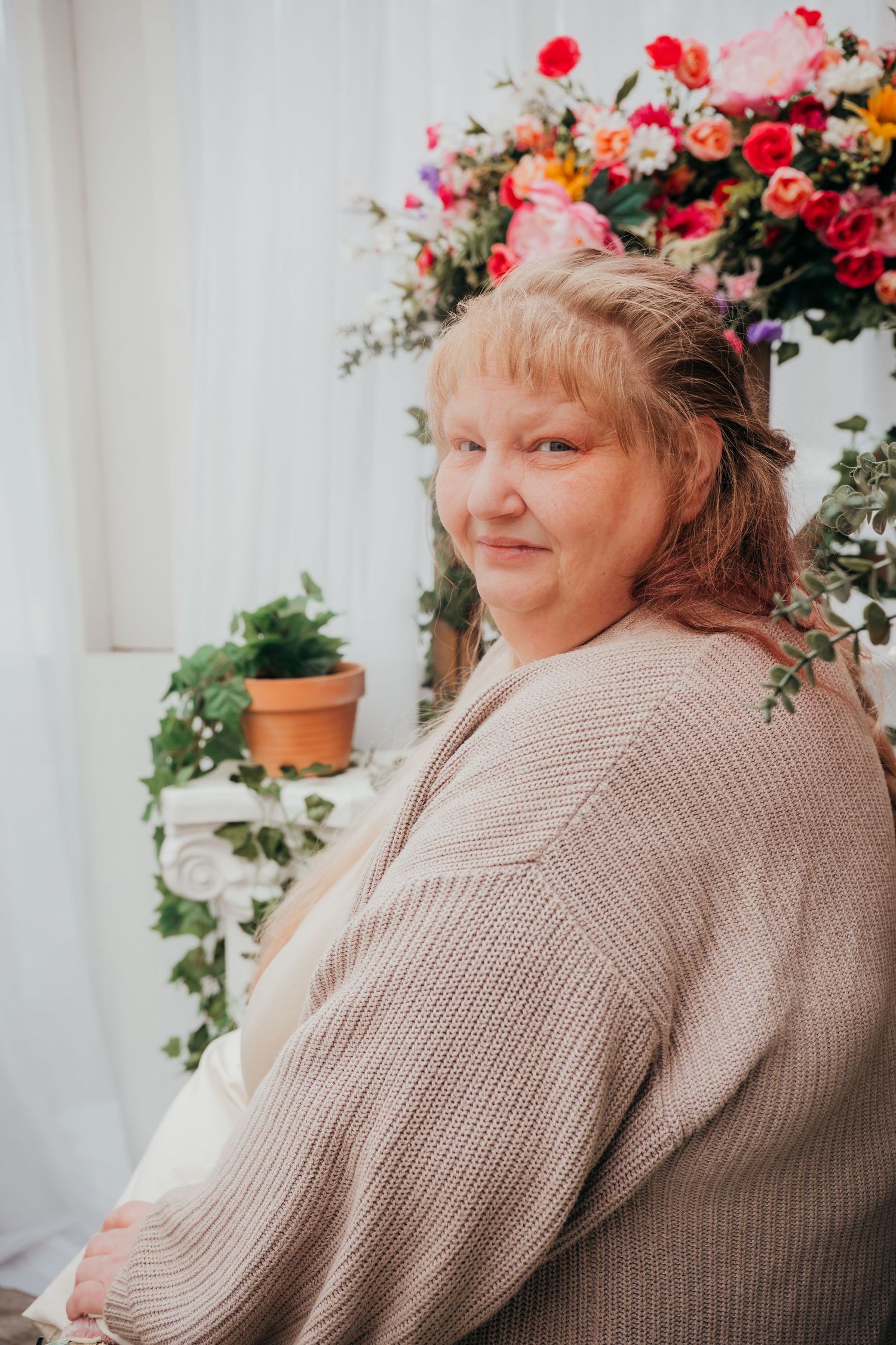 A person wearing a beige sweater sits beside colorful floral arrangements and potted plants in a bright indoor setting.