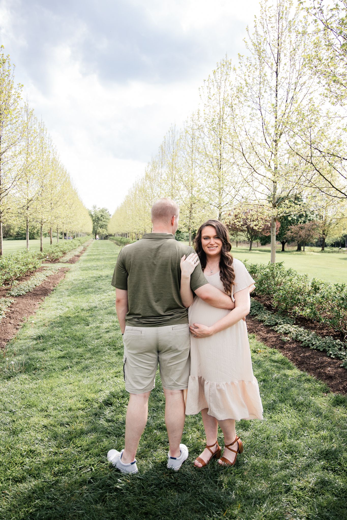 light and airy photo of a pregnant couple standing in between rows of trees smiling.