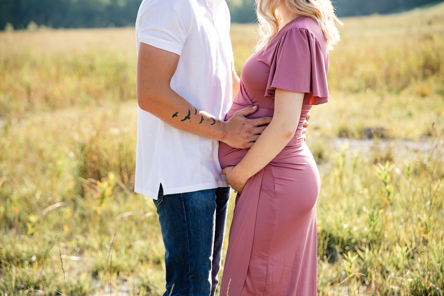 light and airy photo of a pregnant couple standing in a field holding her belly.