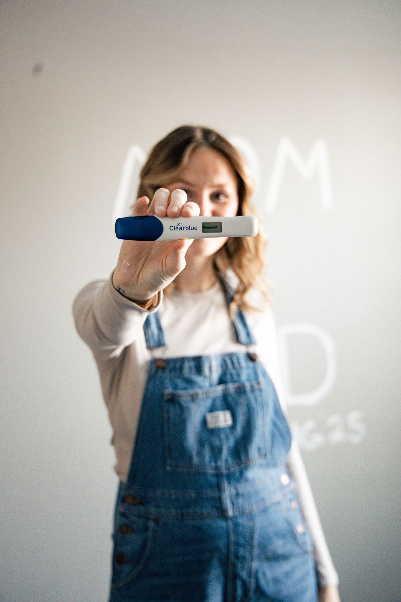 light and airy photo of a woman holding a positive pregnancy test in blue overalls