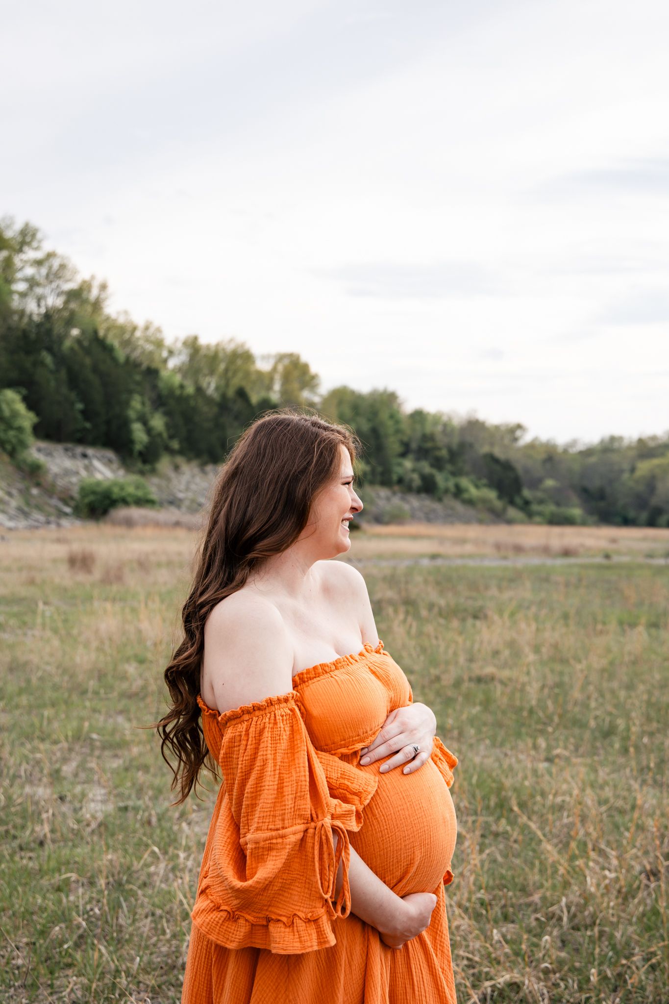 light and airy photo of a pregnant mother in an orange dress looking at her husband laughing in a field.