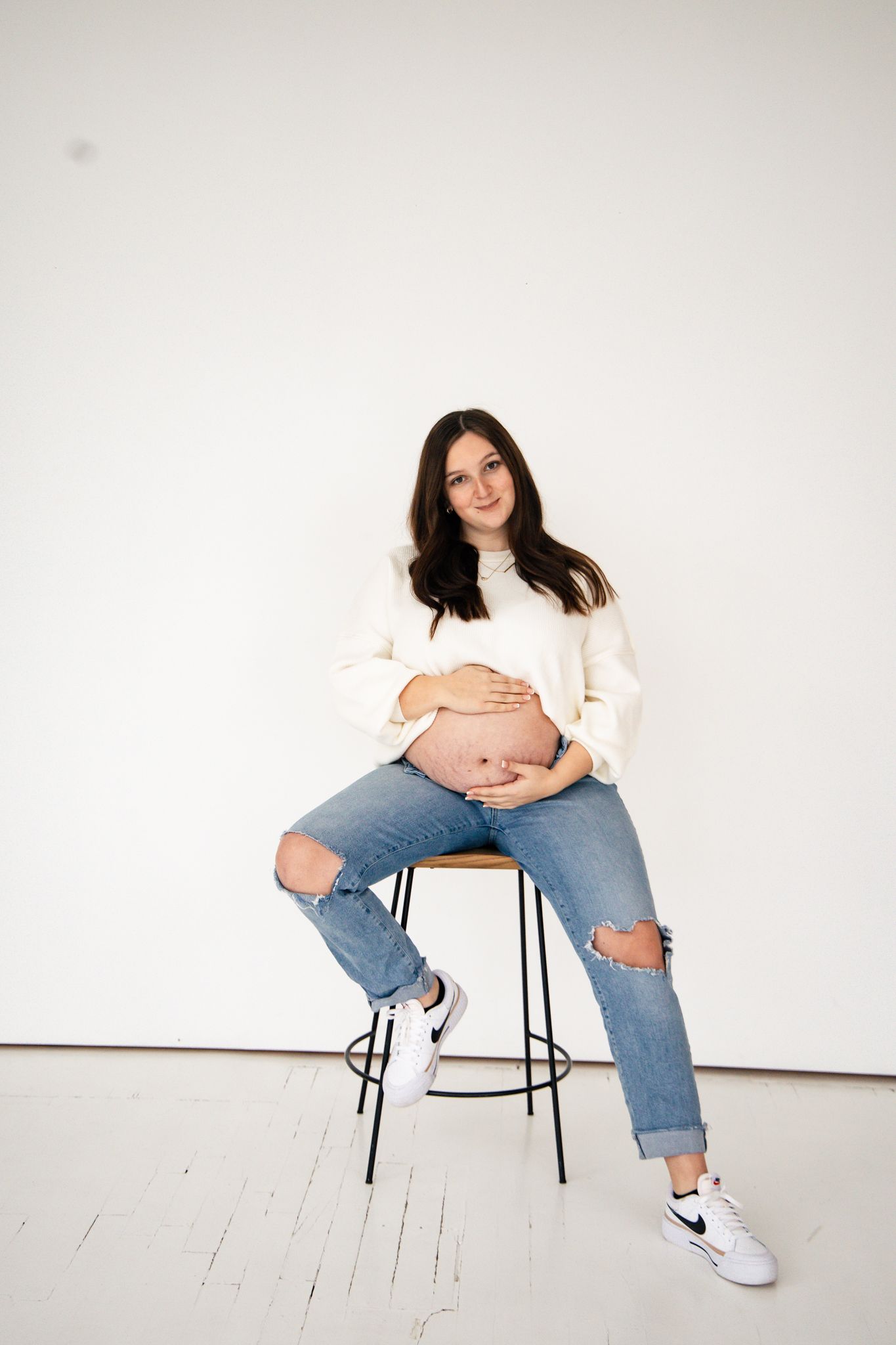 light and airy photo of a pregnant woman sitting on a bench with a white wall looking off to the side smiling.