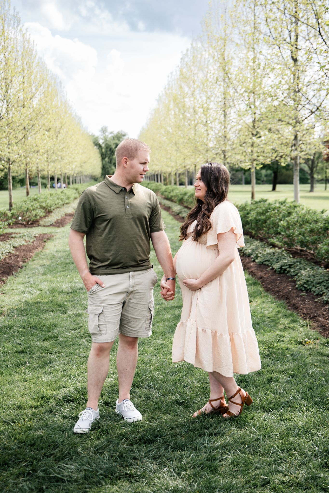 light and airy photo of a couple in between a row of trees smiling, pregnant.
