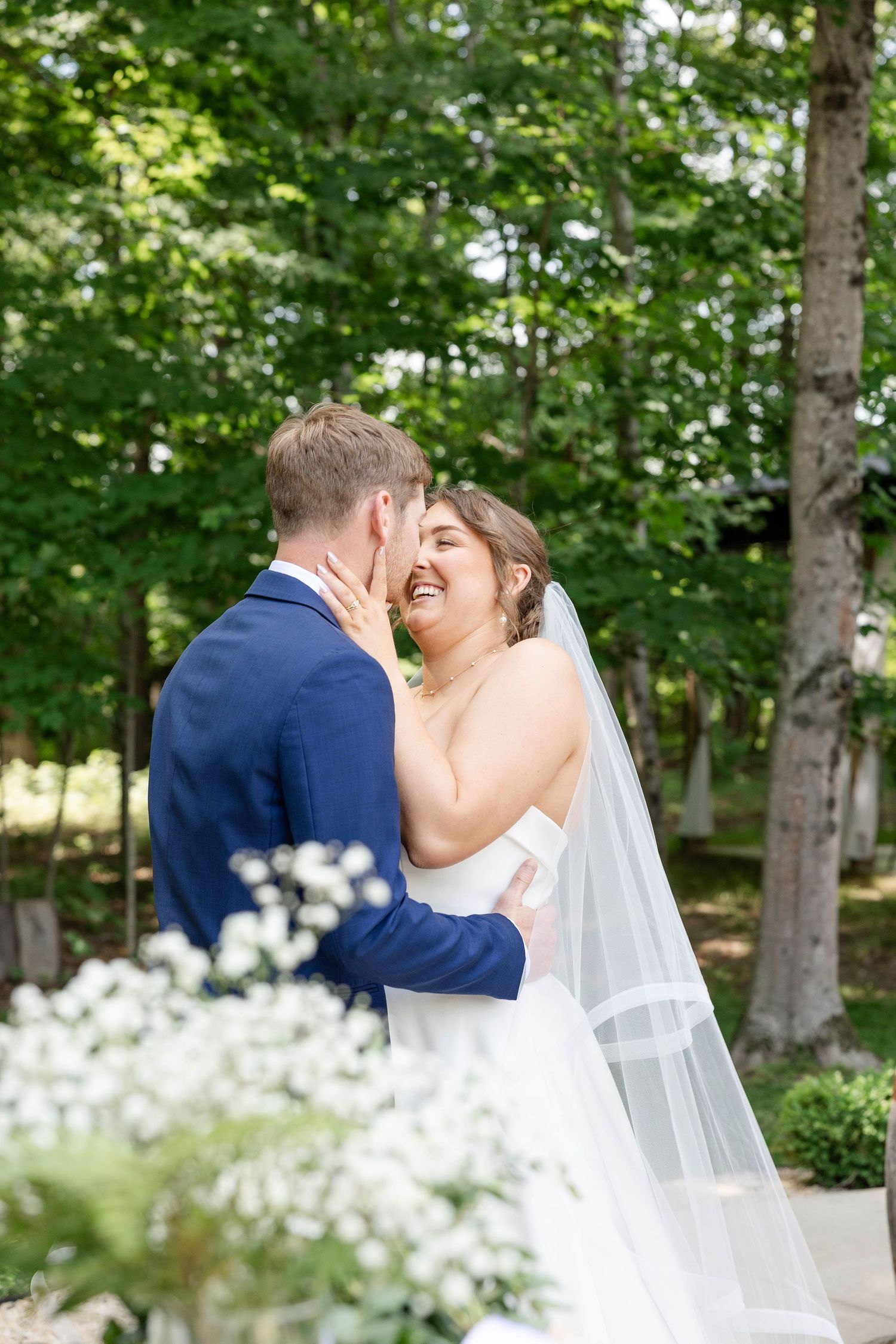 Bride & Groom sharing a romantic embrace outdoors on their wedding day, surrounded by greenery.