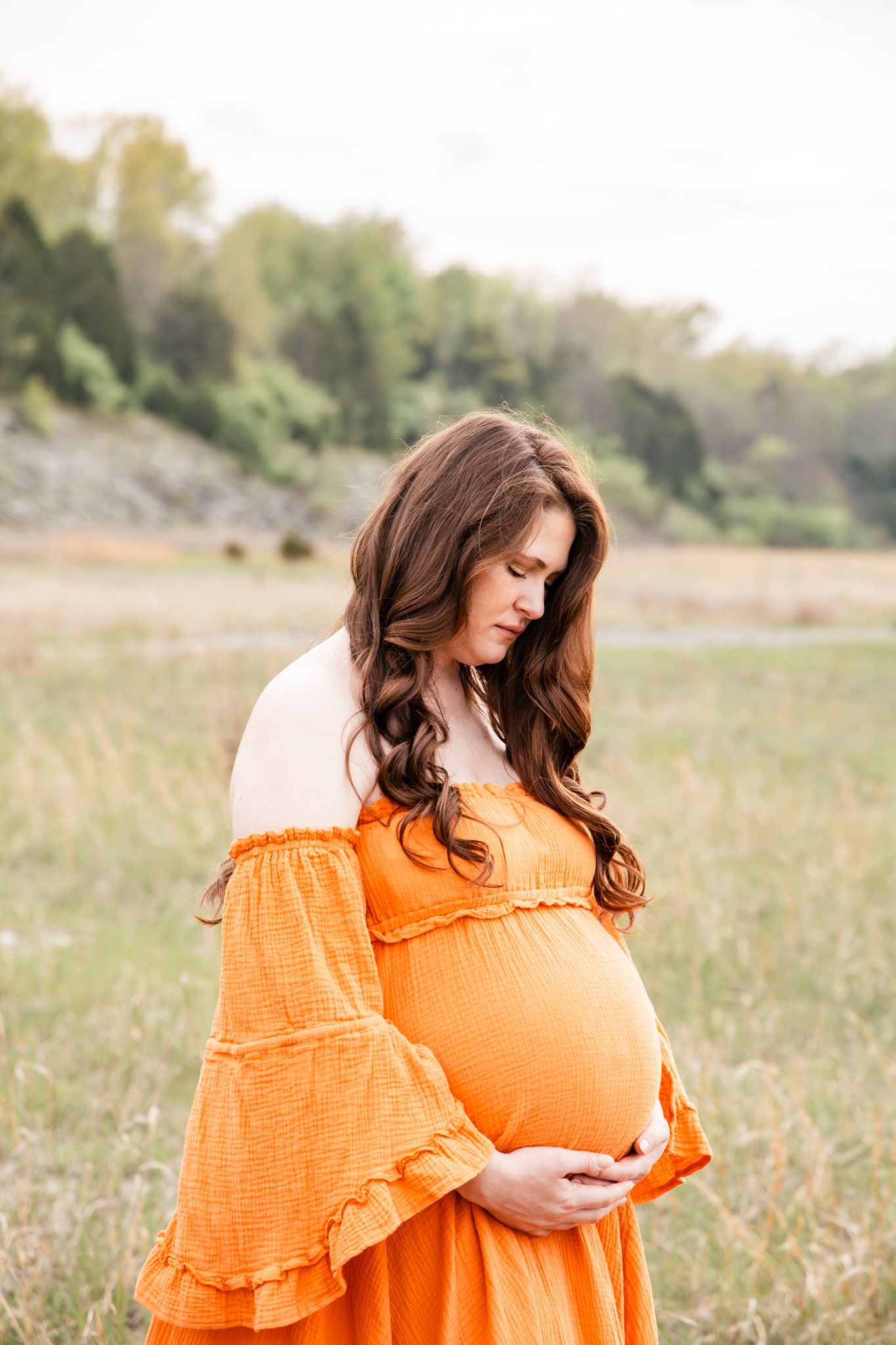 light and airy photo of a pregnant mother in an  orange dress looking down at her bump in a field.