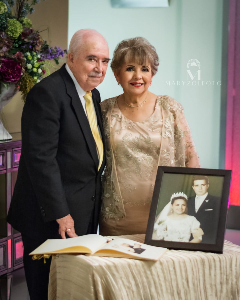 Pareja de adultos mayores vestidos formalmente. Frente ellos hay una mesa con un libro abierto y un portarretrato con una fotografía.