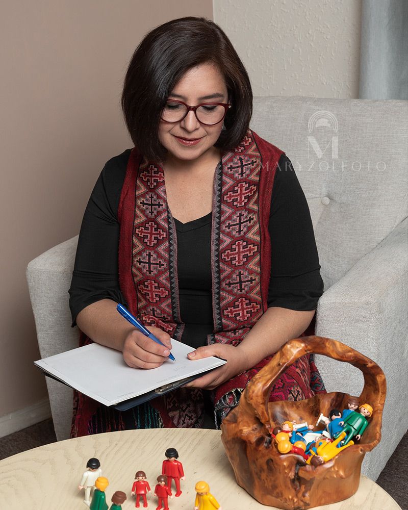 Mujer profesionista sentada tomando apuntes, frente a ella hay una mesa con figuras Playmobil.